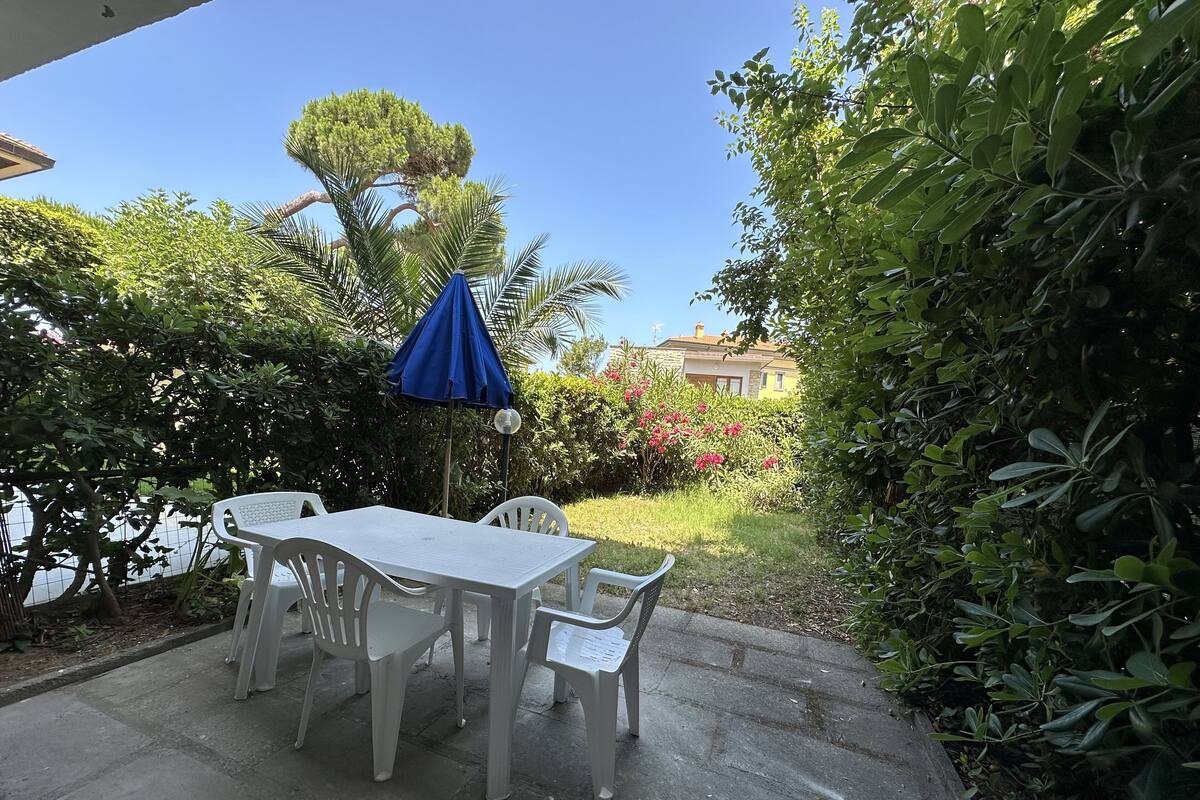 A patio with a table and chairs, a blue umbrella, and lush greenery.