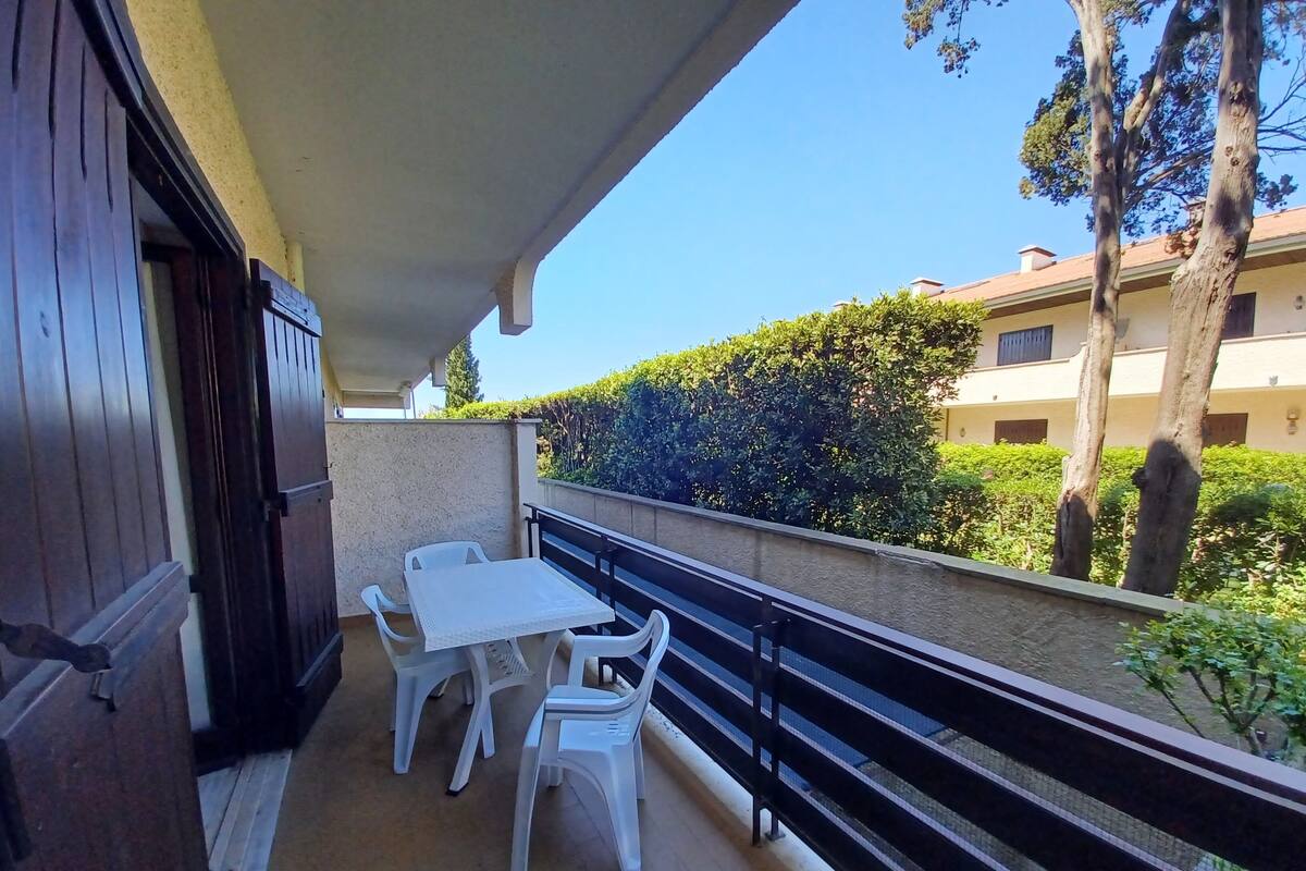 A balcony with a table and chairs, a railing, and a view of a hedge and buildings.