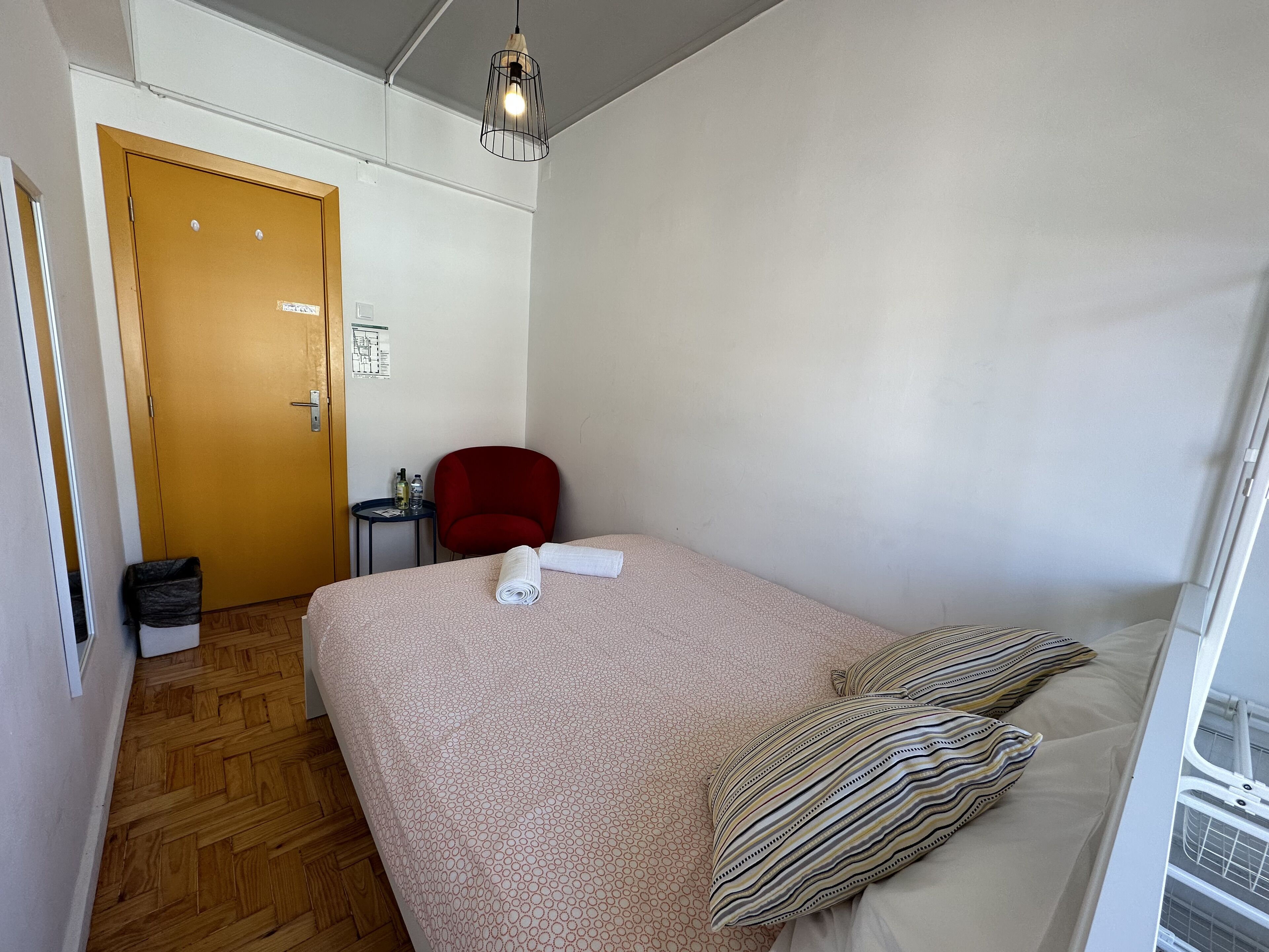 A hotel room with a bed, a red chair, a small table with a bottle and a glass, and a wooden door.