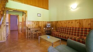 A cozy living room with wooden paneling, a plaid sofa, a television, and a dining area with chairs and a table. - Picu Castiellu Aparthotel (Llanes)