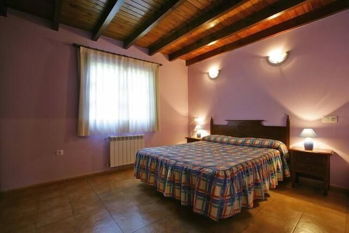 Un dormitorio con techo de madera, una cama con colcha escocesa, dos lámparas de noche y un ventanal con cortinas.