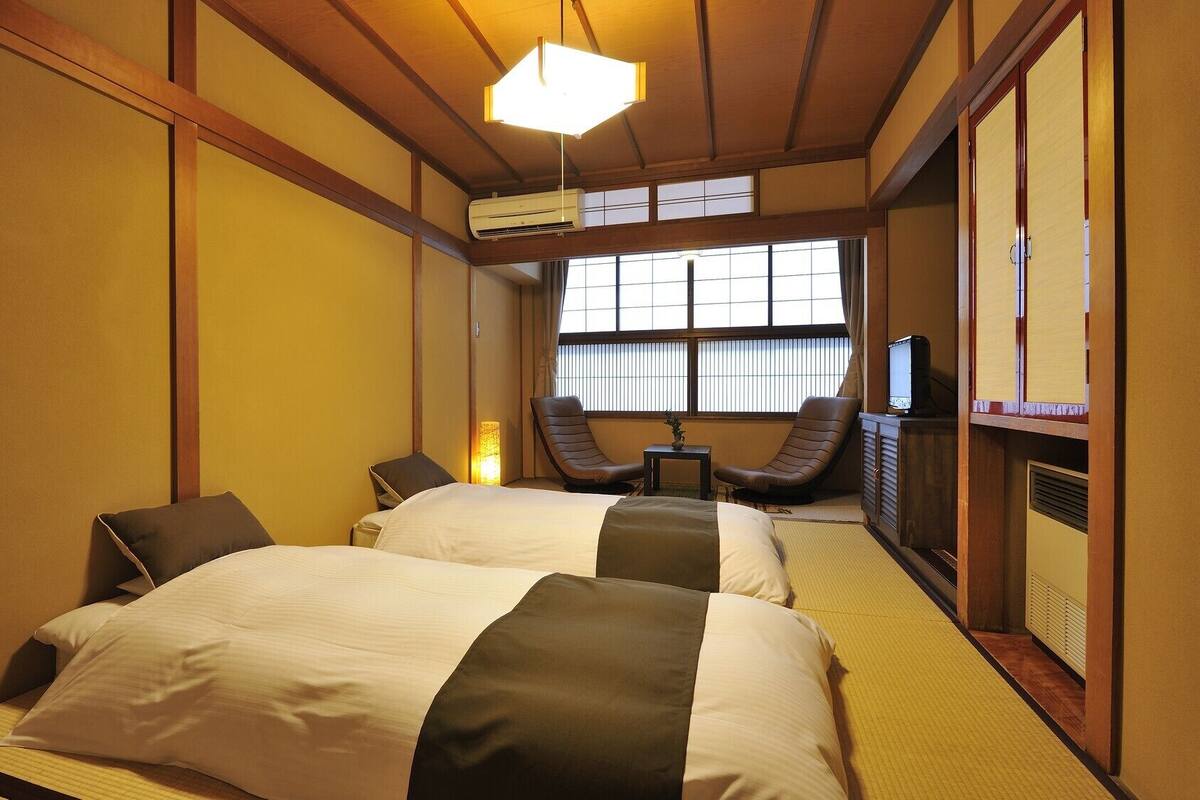 Una habitación de hotel tradicional japonesa con suelo de tatami, una cama con colcha blanca y marrón, un televisor y una puerta corredera.