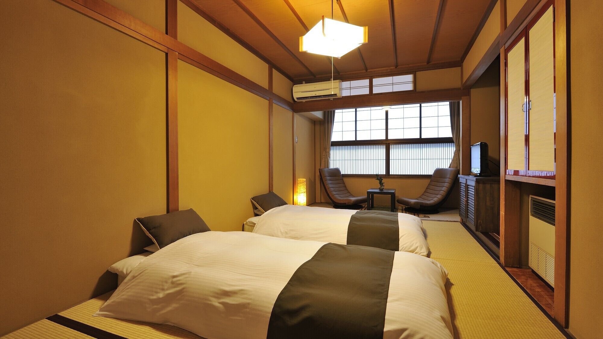 Een traditionele Japanse hotelkamer met tatami vloeren, een bed met een wit en donkerbruin sprei, een televisie en een schuifdeur.