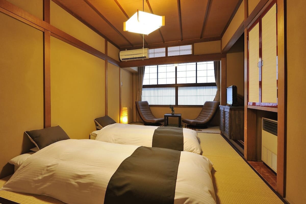 Una habitación de hotel tradicional de estilo japonés, con suelo de tatami, una cama con colcha blanca y marrón oscuro, un televisor y una puerta corredera.