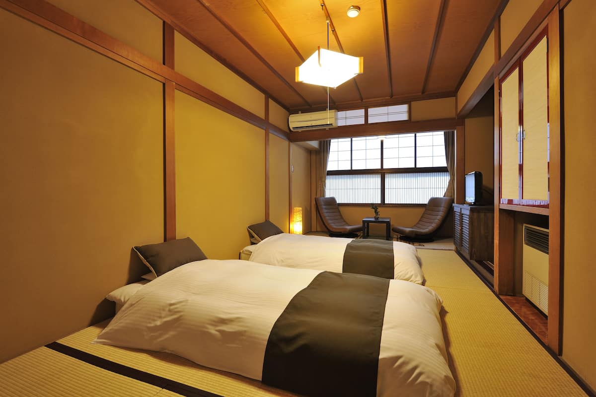 Habitación de hotel tradicional al estilo japonés, con suelo de tatami, dos camas con ropa de cama blanca y oscura, un ventilador de techo y un televisor.