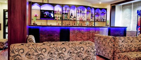 A bar area with a brick wall, shelves of liquor bottles, and a flat-screen TV. There are patterned sofas in the foreground.