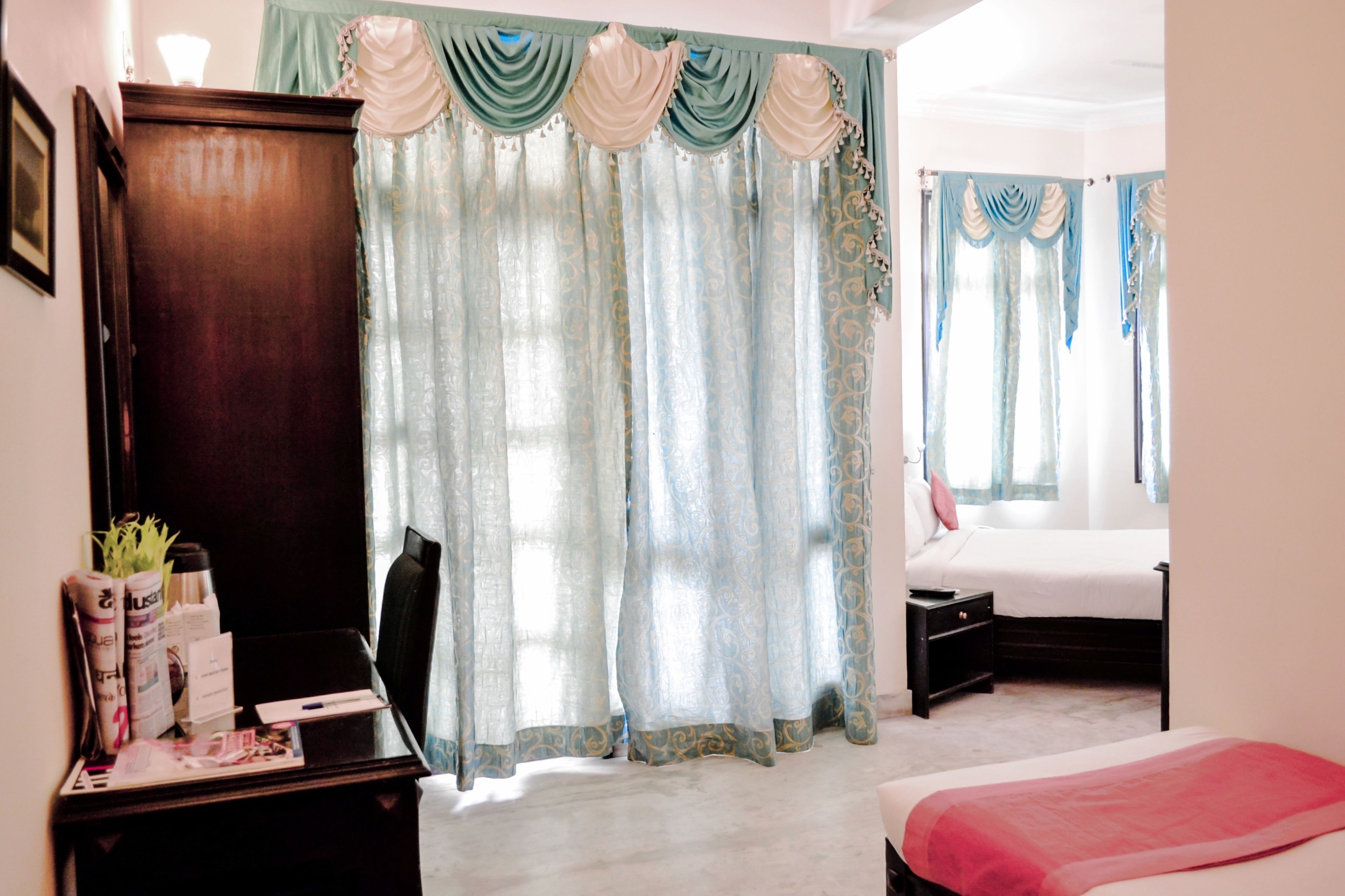 A hotel room with a wooden dresser, a desk with a lamp, a bed with a red blanket, and light blue curtains with a decorative trim.