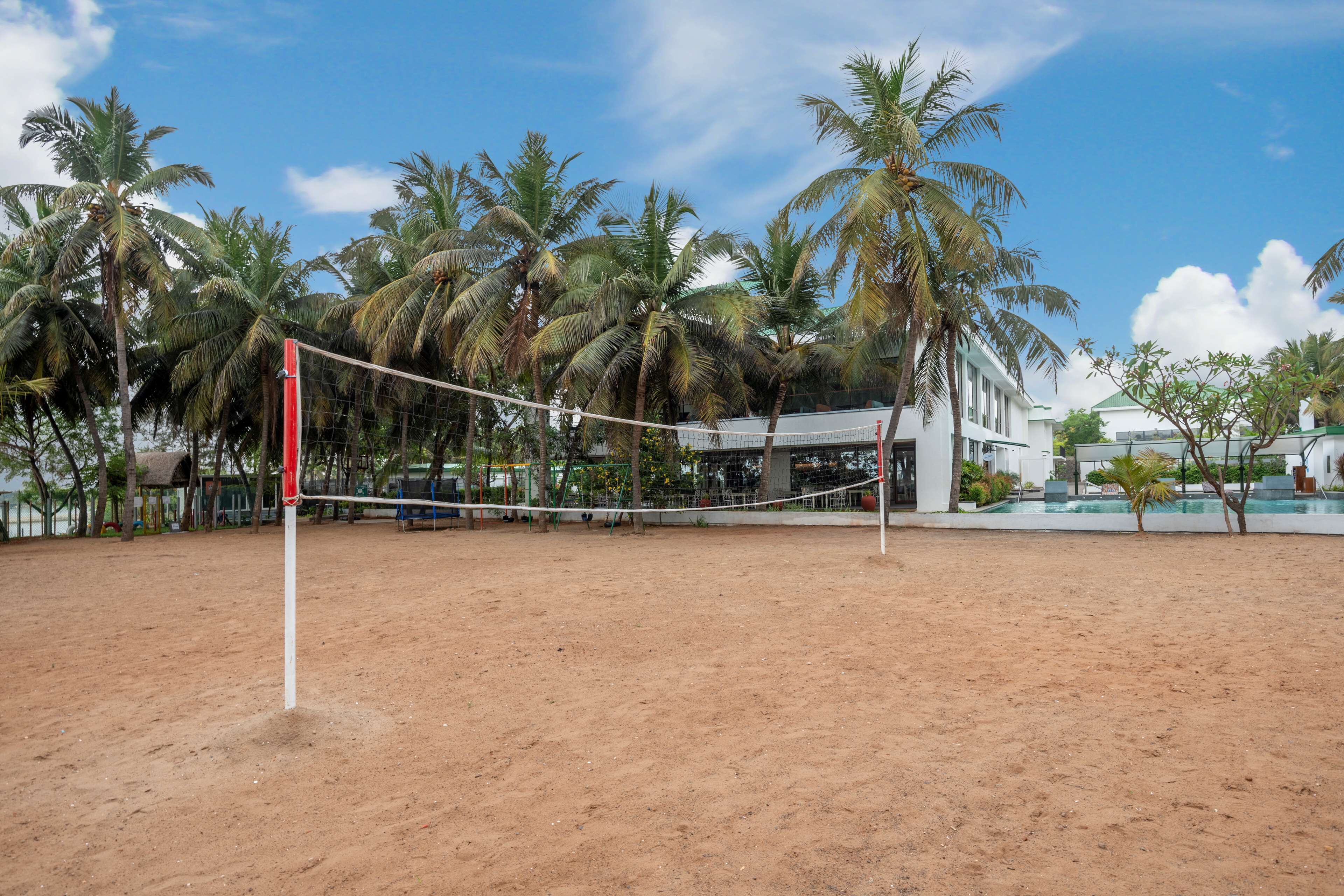 A sandy beach volleyball court with a net, surrounded by palm trees and a building with a green roof in the background.