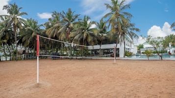 A sandy beach volleyball court with a net, surrounded by palm trees and a building with a green roof in the background.