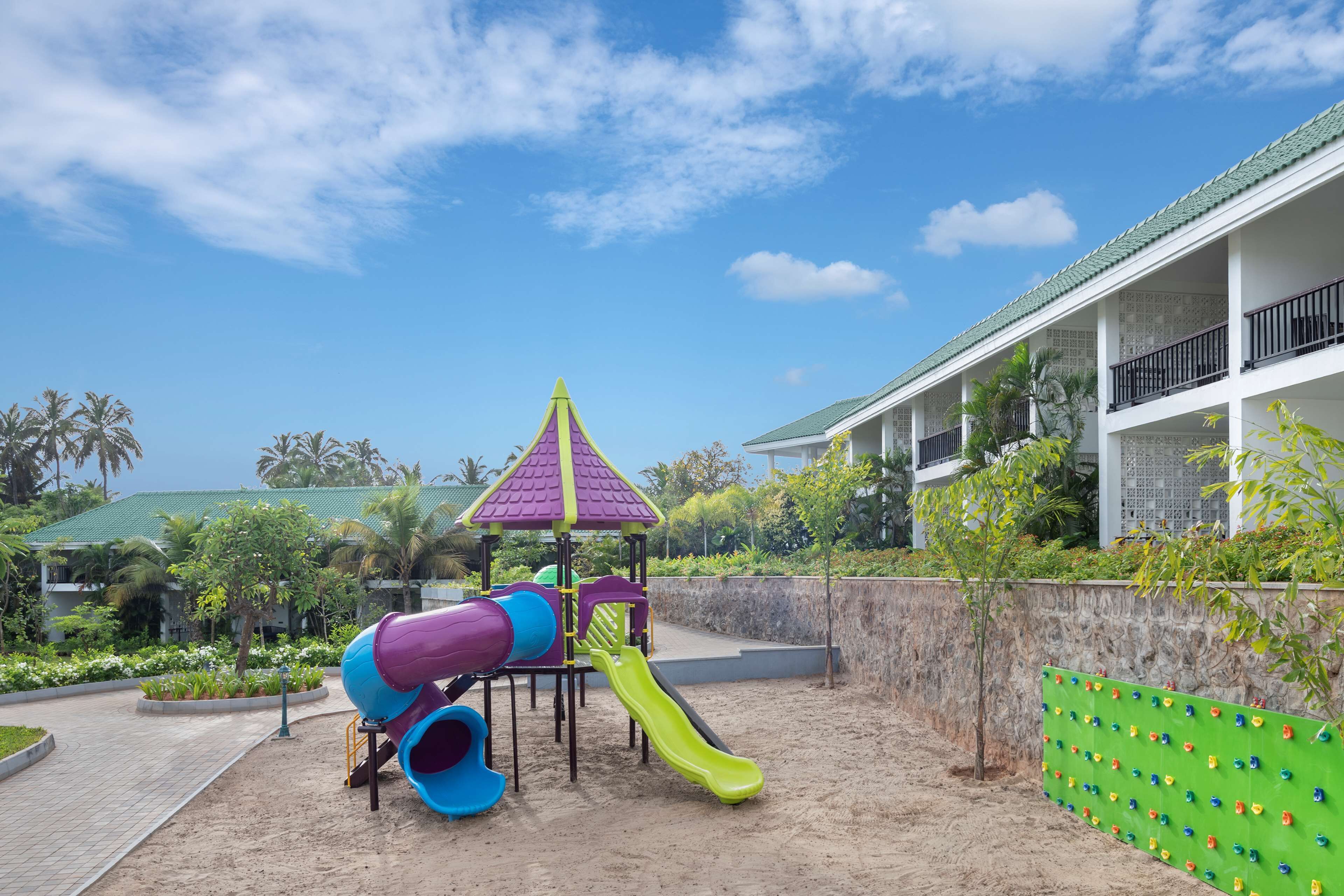 A playground with slides and climbing structures, surrounded by a landscaped garden and a building with a green roof.
