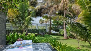 Un jardín tropical con una hamaca, una mesa con mantel estampado y una pequeña construcción de ladrillo.