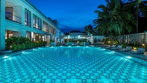 Una piscina grande y bien iluminada con un diseño a cuadros, rodeada de palmeras y un edificio moderno con amplias ventanas.