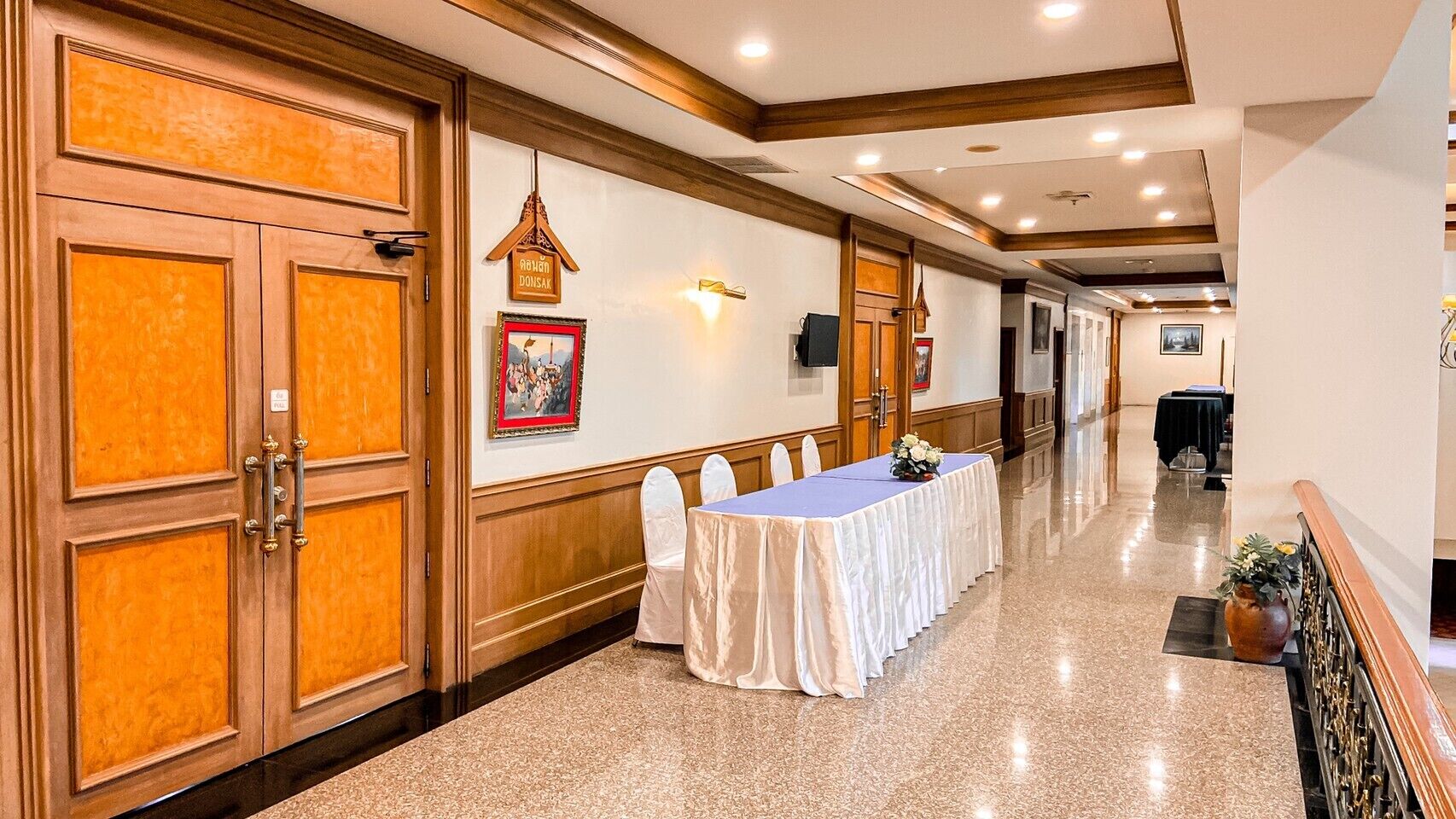 Un corridoio d'hotel con un lungo tavolo apparecchiato per la cena, porte in legno e un quadro appeso al muro.