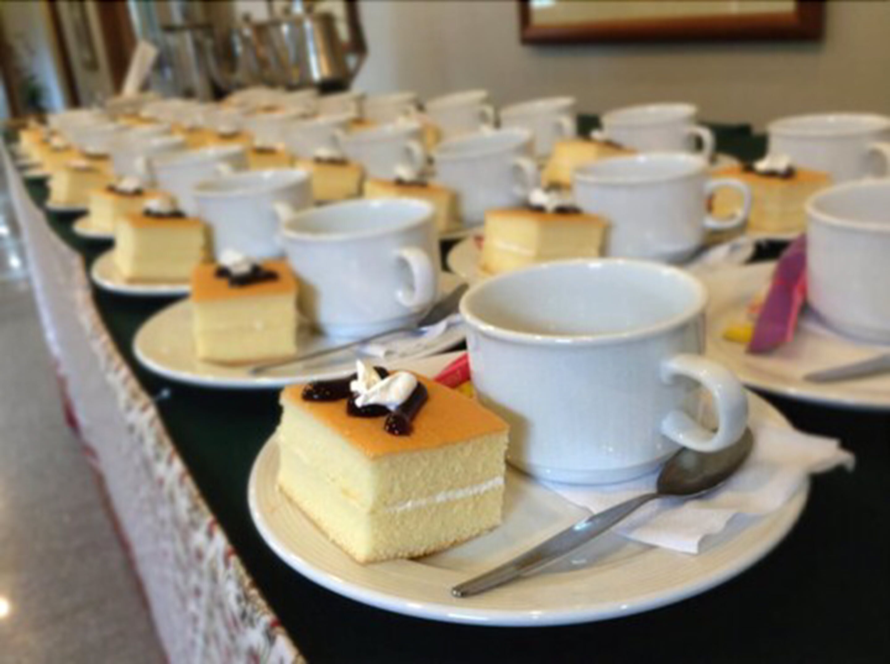Una mesa de buffet con múltiples tazas y platos blancos, cada uno con una porción de pastel y una cuchara.