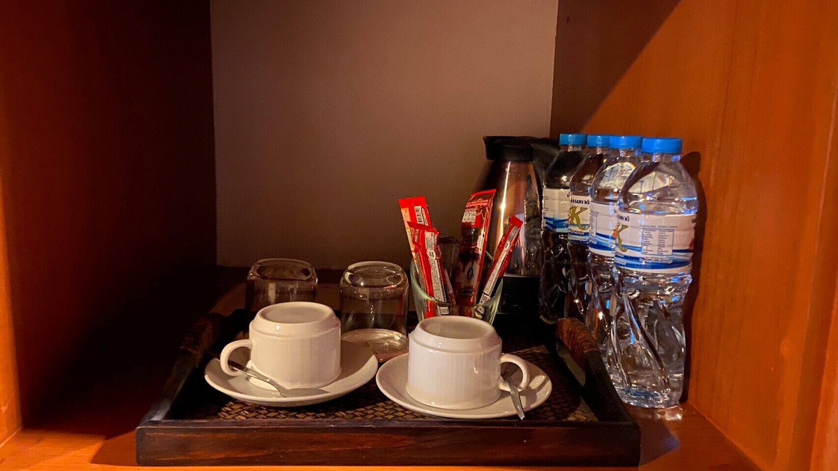 Una bandeja con dos tazas y platillos blancos, un vaso, una botella de agua y paquetes de bocadillos.