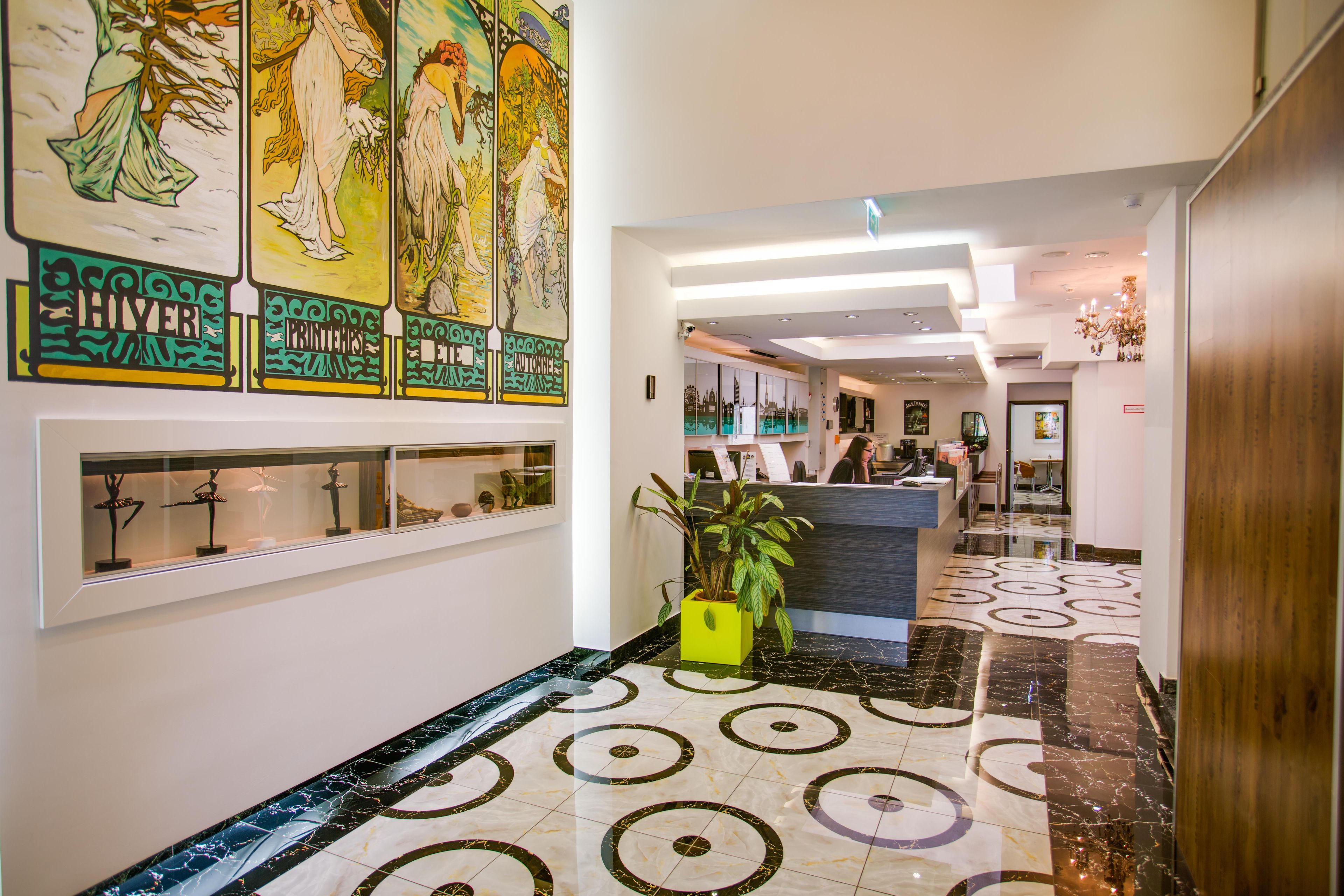 A hotel lobby with a reception desk, a large art piece on the wall, and a patterned floor.