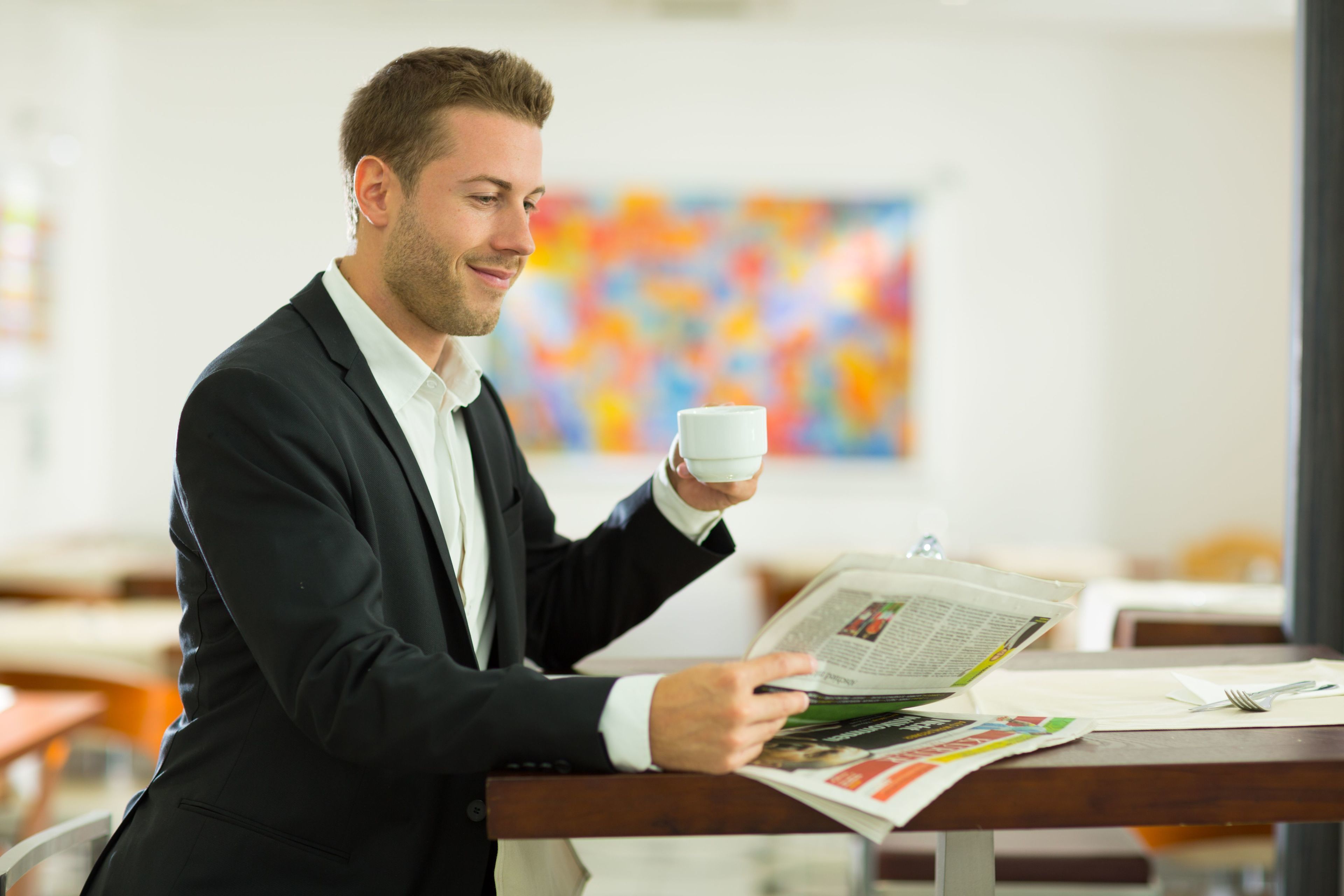 A man in a suit reading a newspaper at a wooden table in a room with a colorful abstract painting on the wall.
