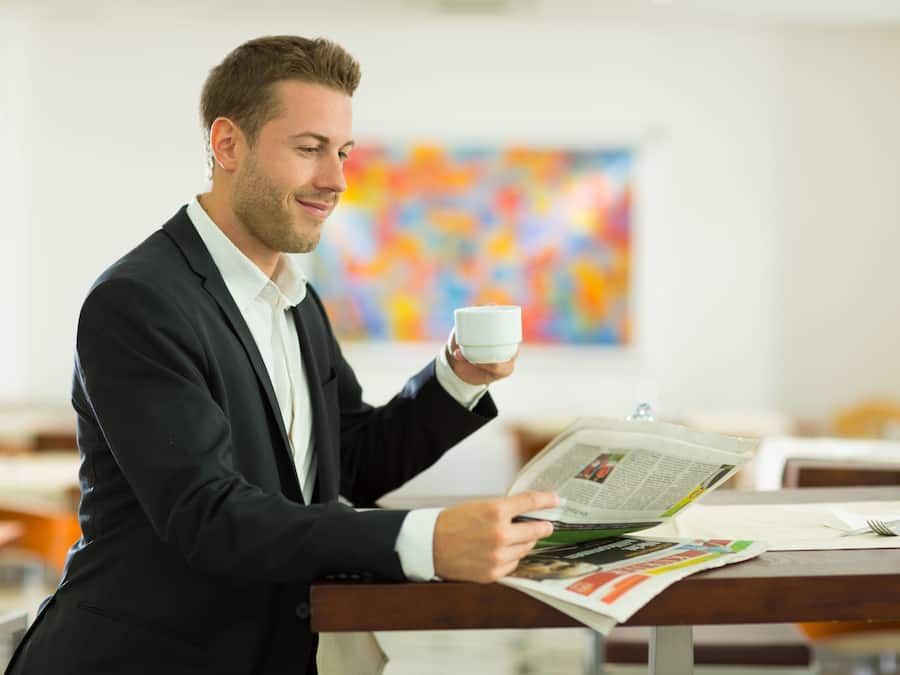 Un hombre con traje lee un periódico en una mesa de madera en una habitación con un cuadro abstracto y colorido en la pared.