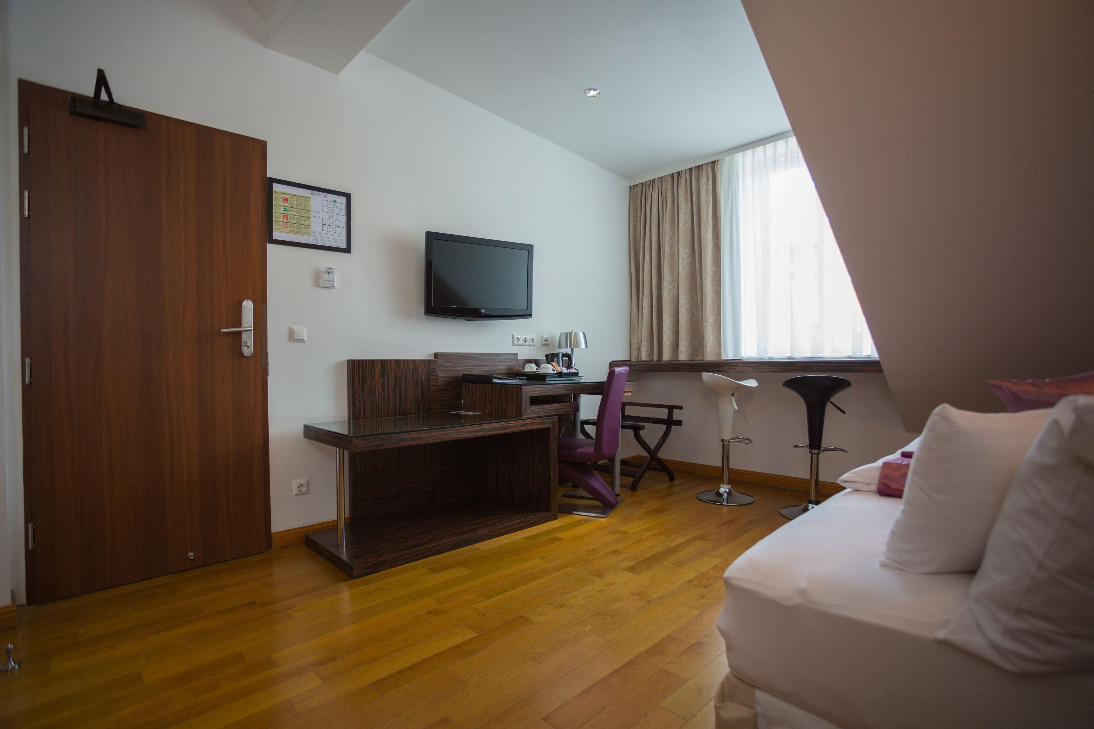 Una habitación de hotel moderna con suelo de madera, un televisor de pantalla plana montado en la pared, un escritorio con una silla, un sofá y una ventana con cortinas.
