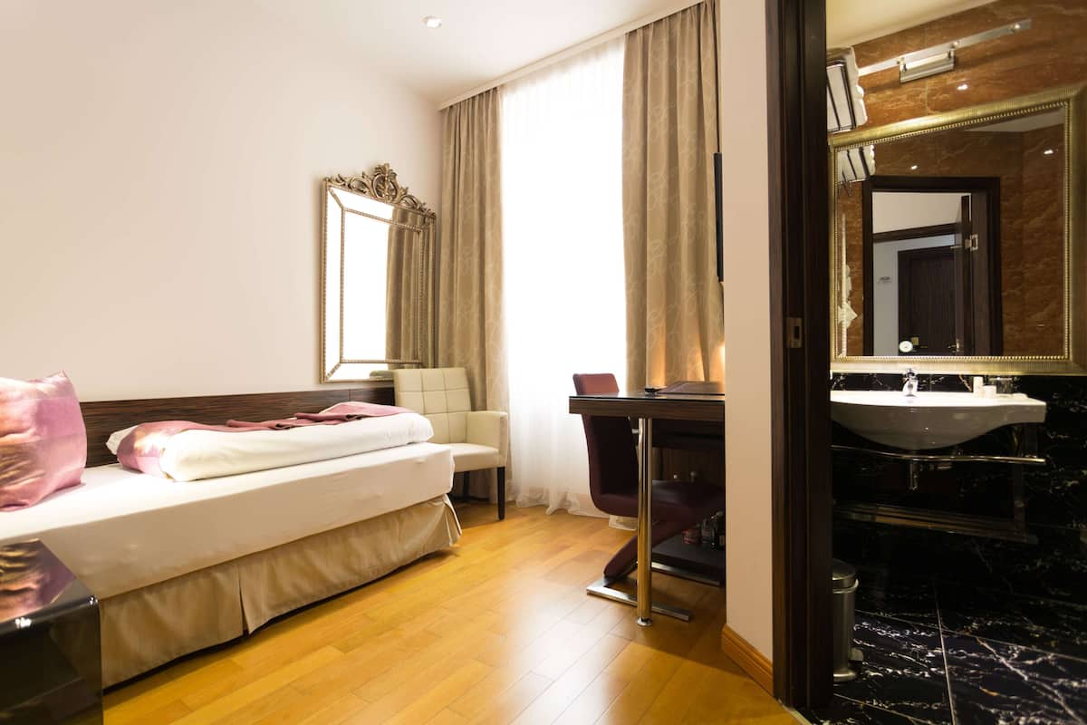 A hotel room with a bed, a desk, a chair, a mirror, and a bathroom with a sink and mirror.