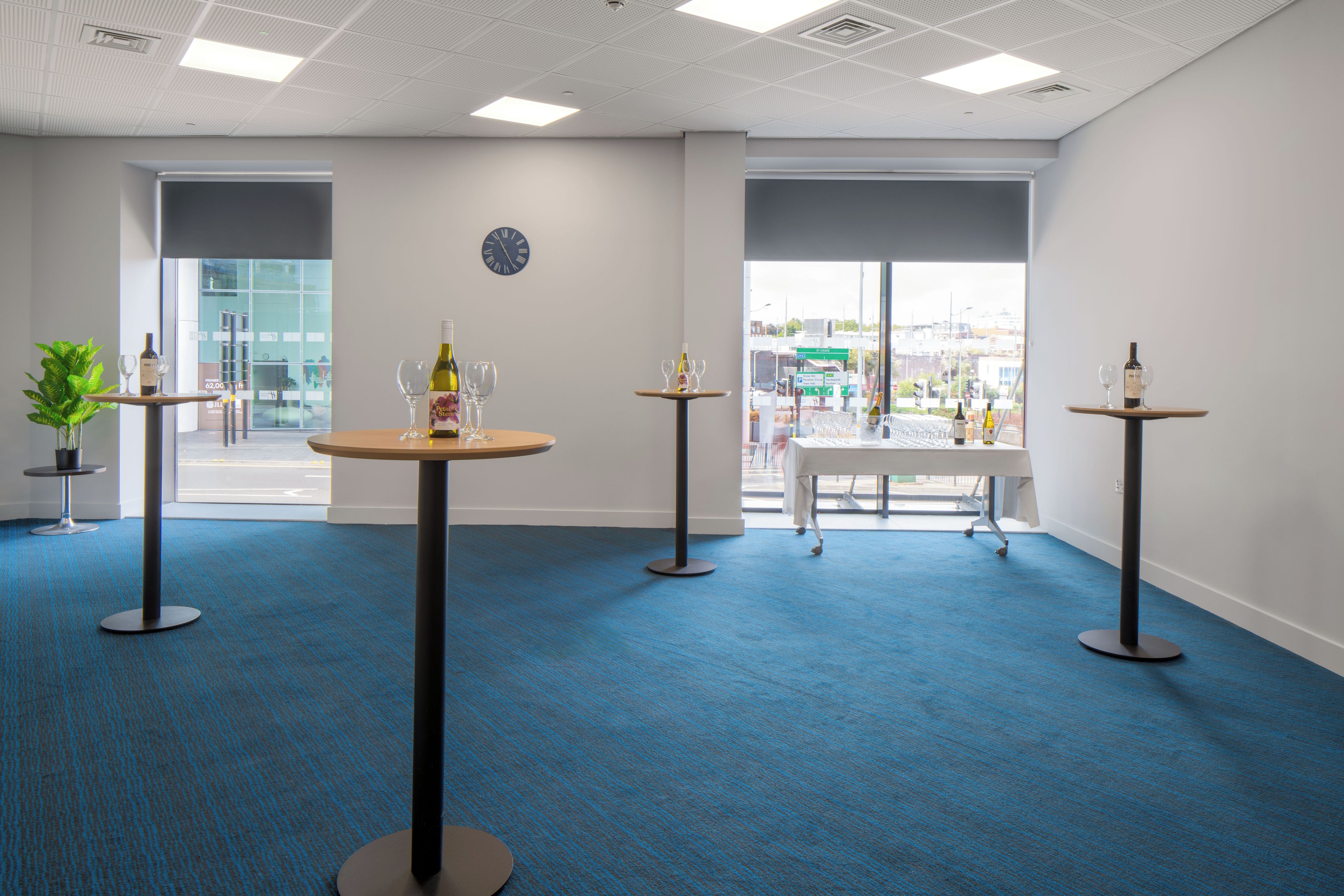 A modern conference room with blue carpeting, multiple tables with wine bottles and glasses, a clock on the wall, and large windows offering a view of the city.