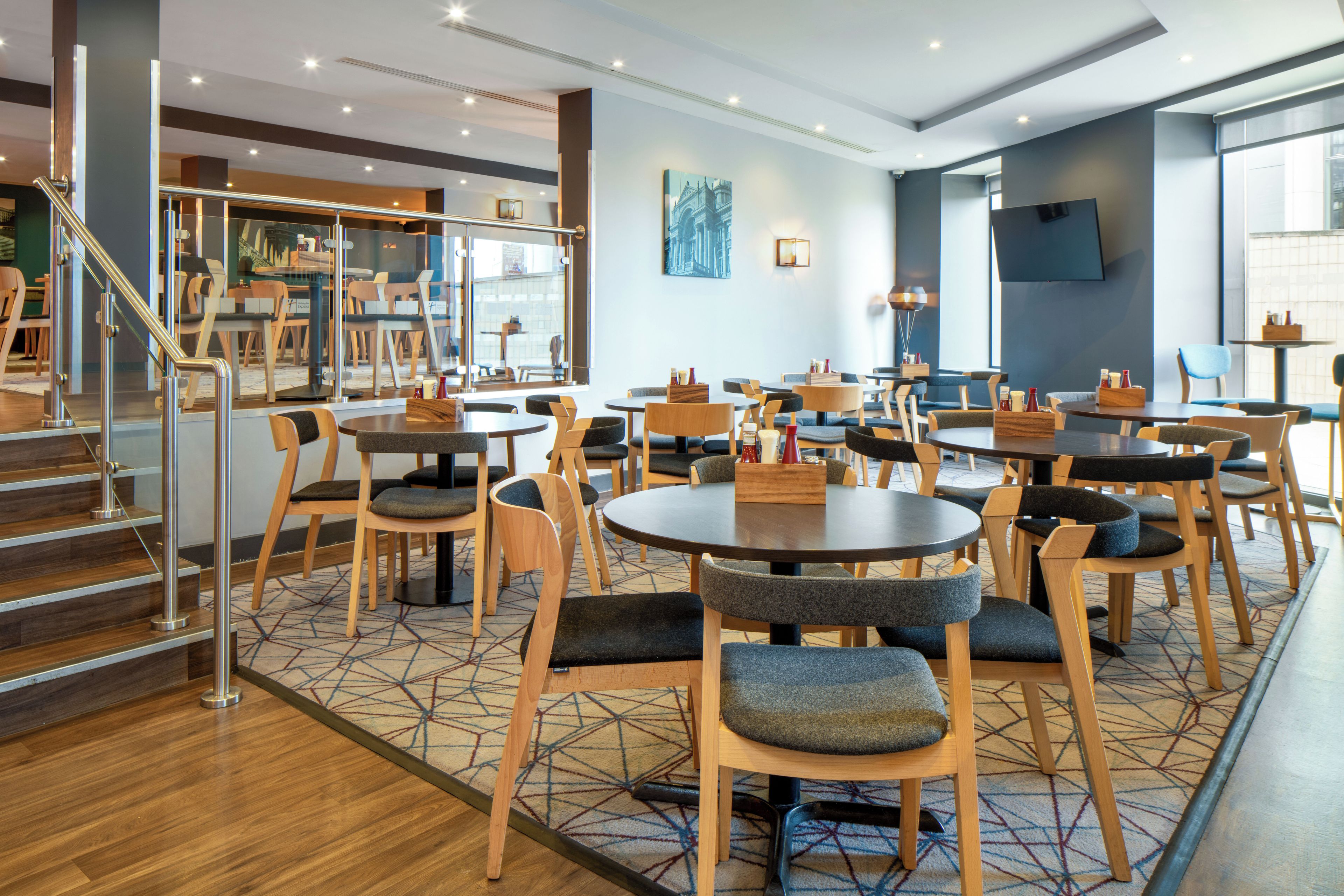 Comedor de hotel con varias mesas y sillas redondas, una escalera y un ventanal grande.