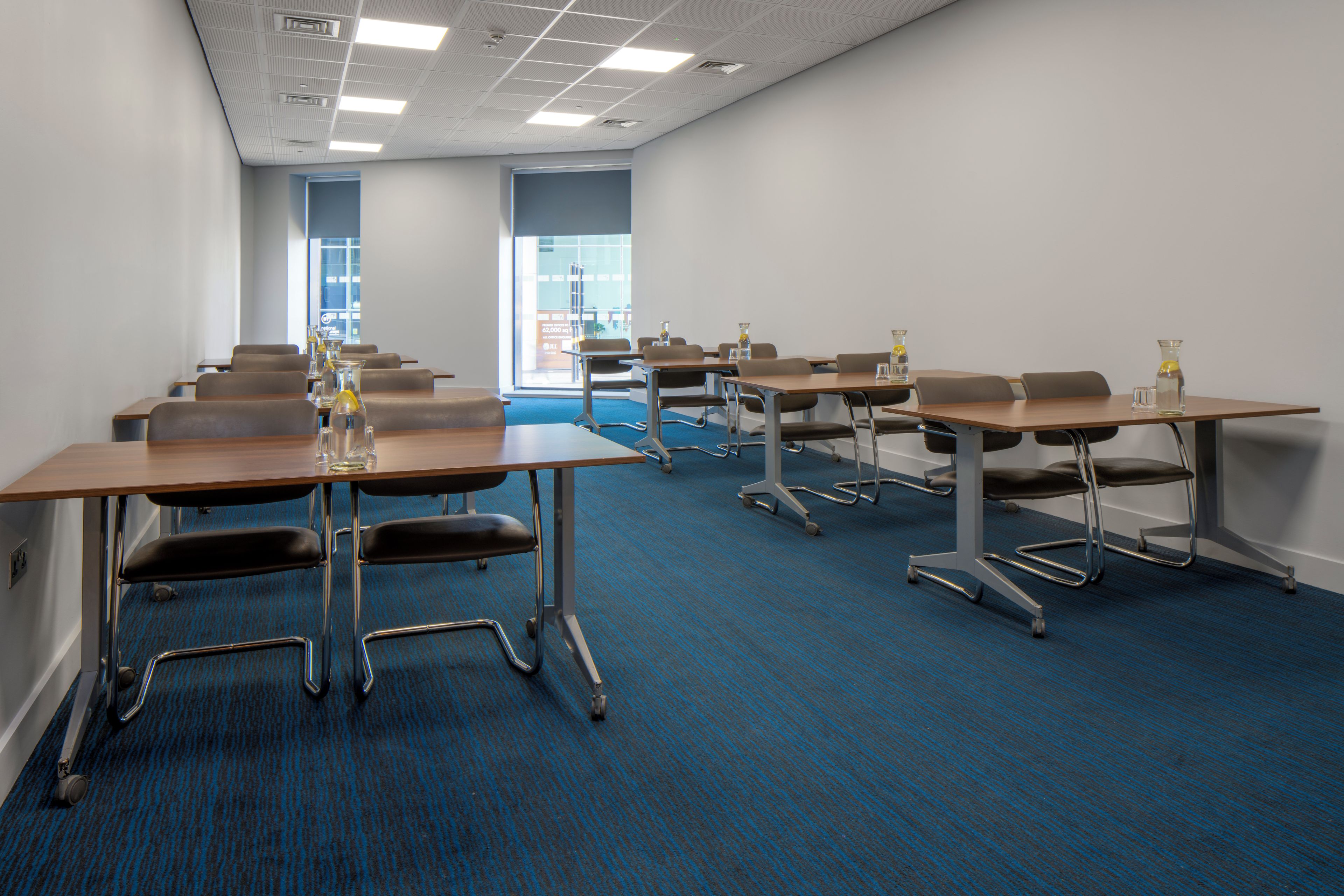 A conference room with multiple tables and chairs, a blue carpet, and a view of the outside through large windows.