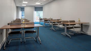 A conference room with multiple tables and chairs, a blue carpet, and a view of the outside through large windows.