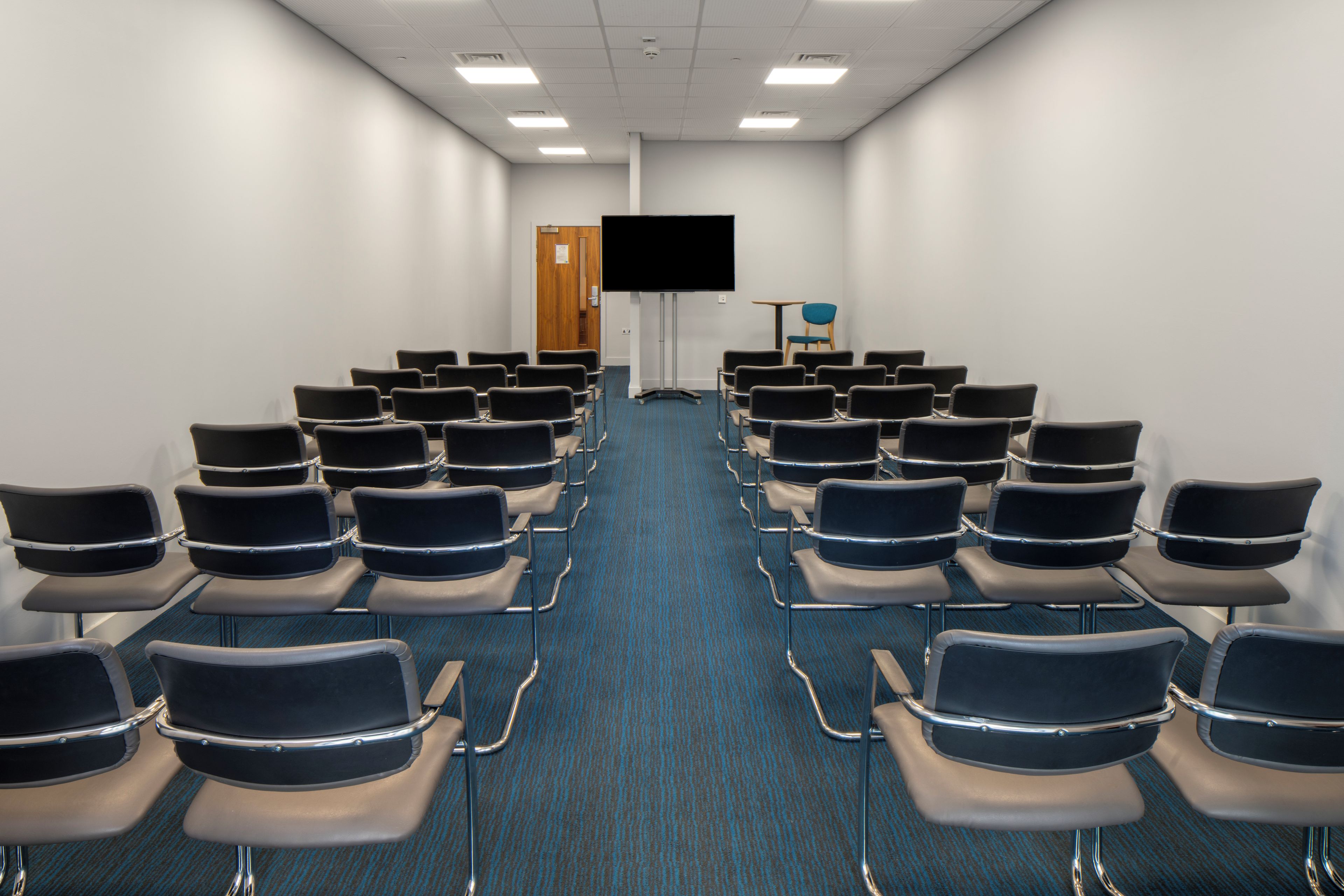 A conference room with rows of chairs facing a black screen, a wooden door, and a small table with a chair.