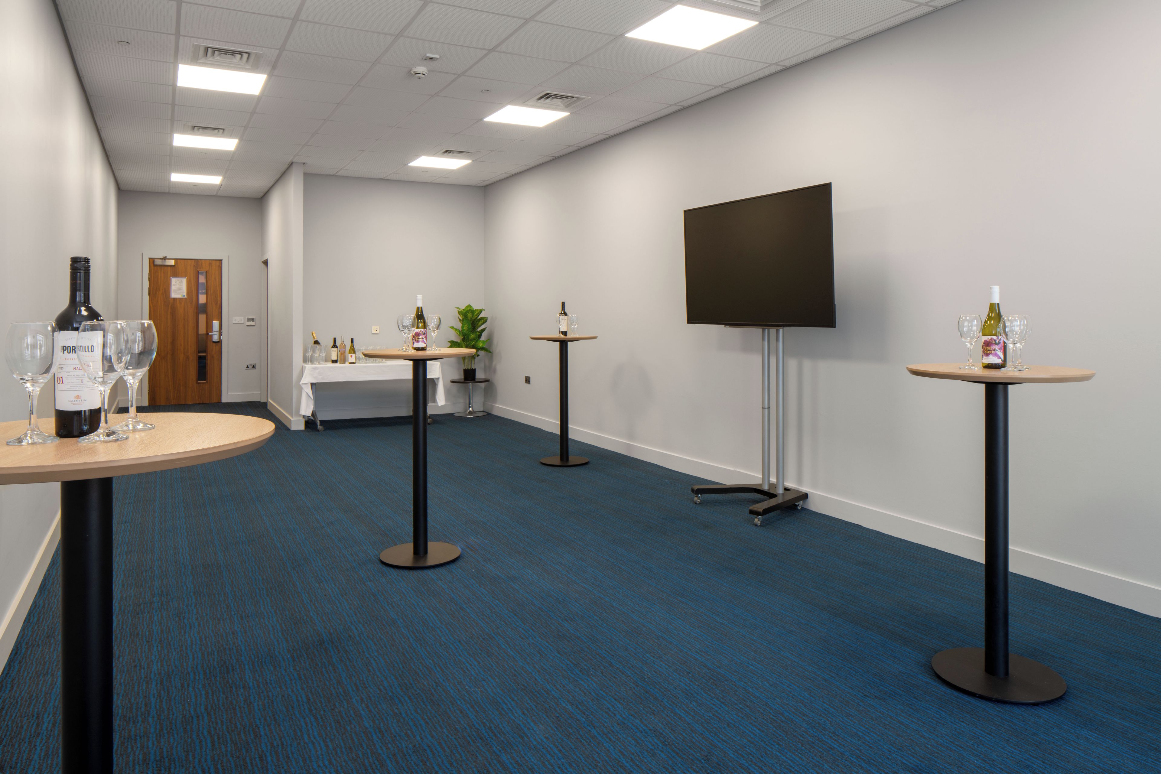 A conference room with a large flat-screen TV, multiple tables with wine bottles and glasses, and a carpeted floor.