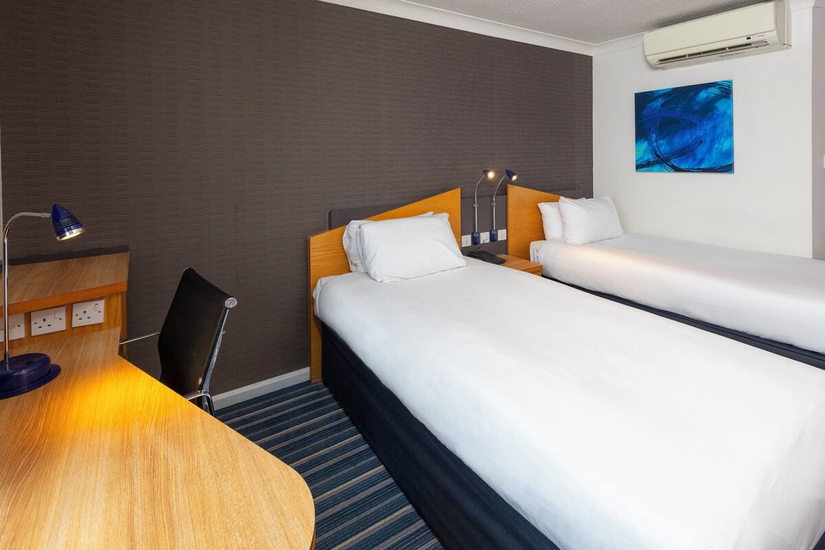 A hotel room with two beds, a desk with a lamp, and an air conditioning unit on the wall.