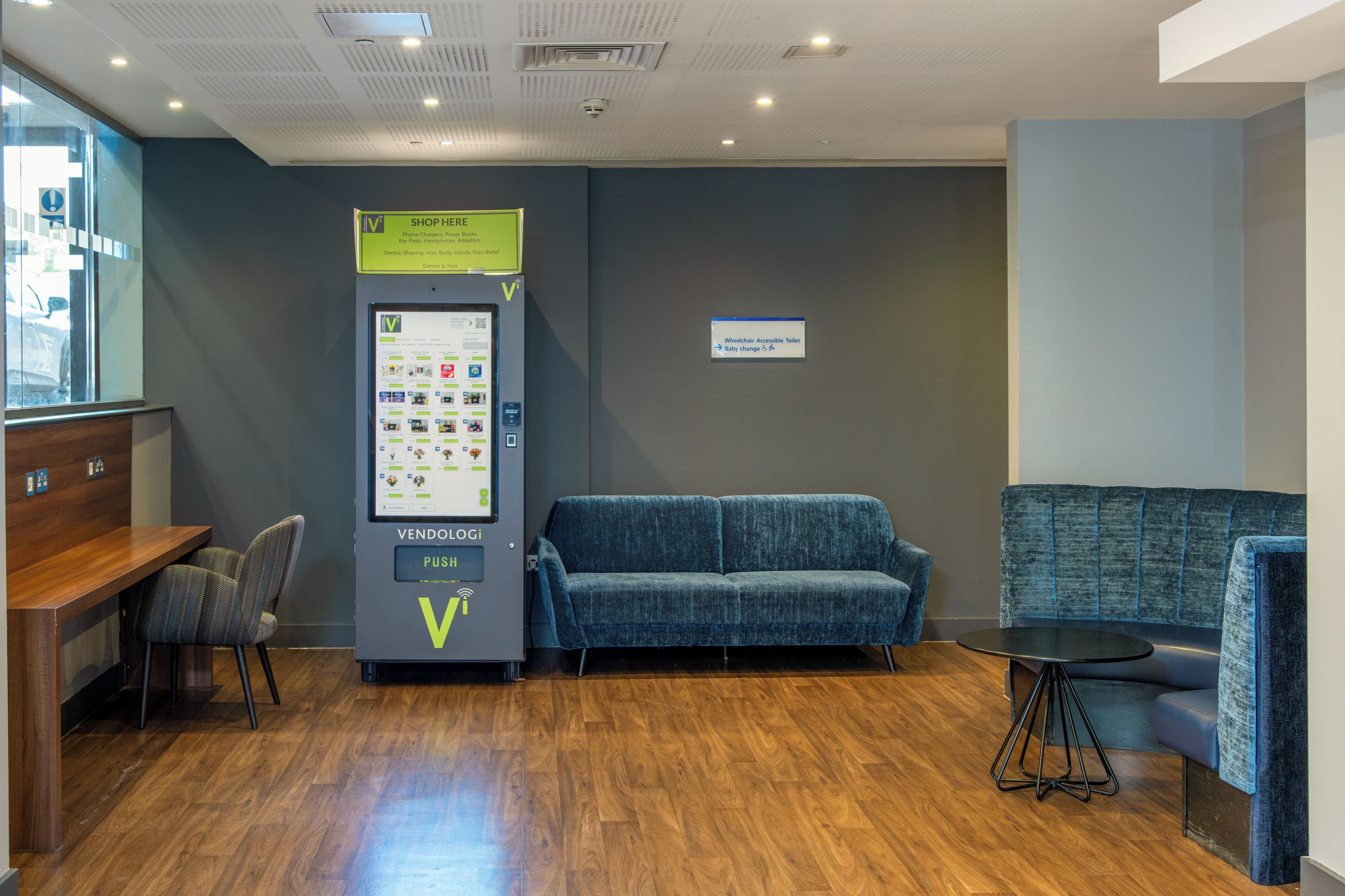 A vending machine with a Venn diagram logo, a sign for a wheelchair accessible toilet, and a blue sofa in a room with wooden flooring.