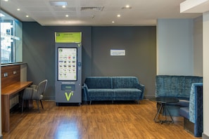 A vending machine with a Venn diagram logo, a sign for a wheelchair accessible toilet, and a blue sofa in a room with wooden flooring. - Holiday Inn Express Birmingham - Snow Hill by IHG (Birmingham)