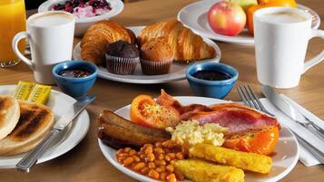 Una mesa de desayuno con platos de comida, incluyendo huevos, tocino, frijoles, salchichas, tostadas y bollería. También hay tazas de café y jugo.