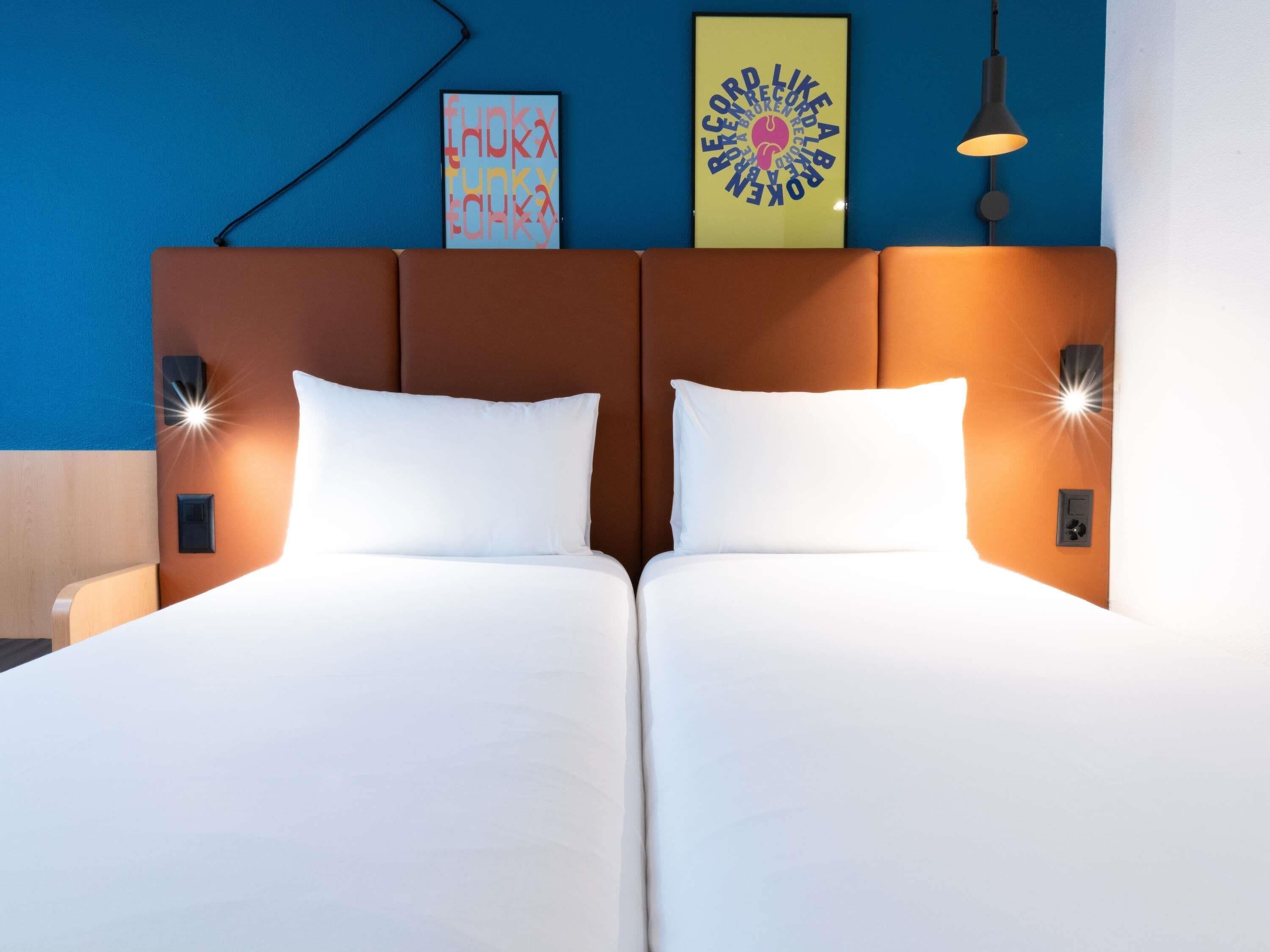 A double bed with white linens, two bedside tables with lamps, a wall-mounted light fixture, and two framed artworks above the bed.