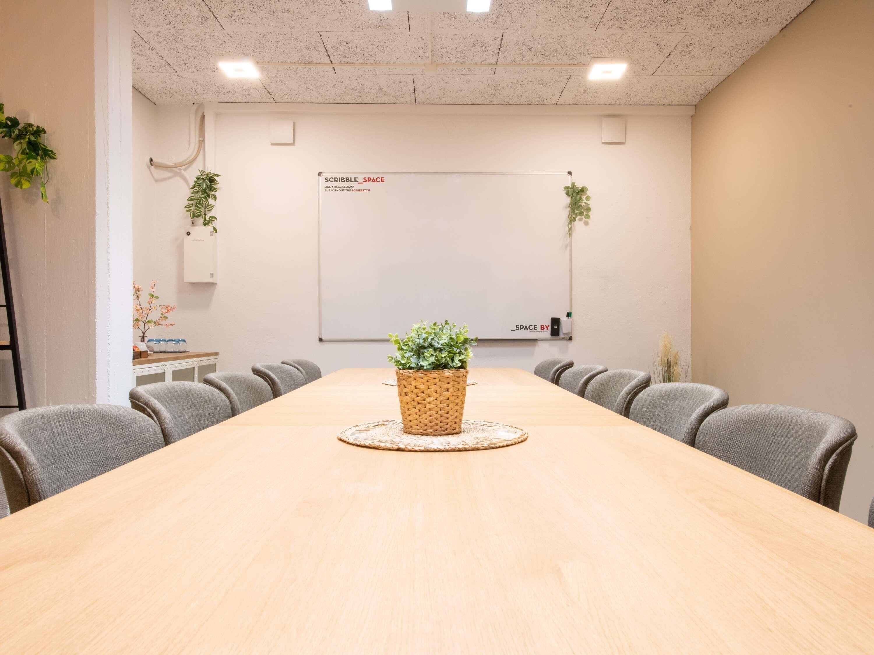 Un salón de conferencias con una larga mesa, sillas grises, una pizarra y plantas en macetas.
