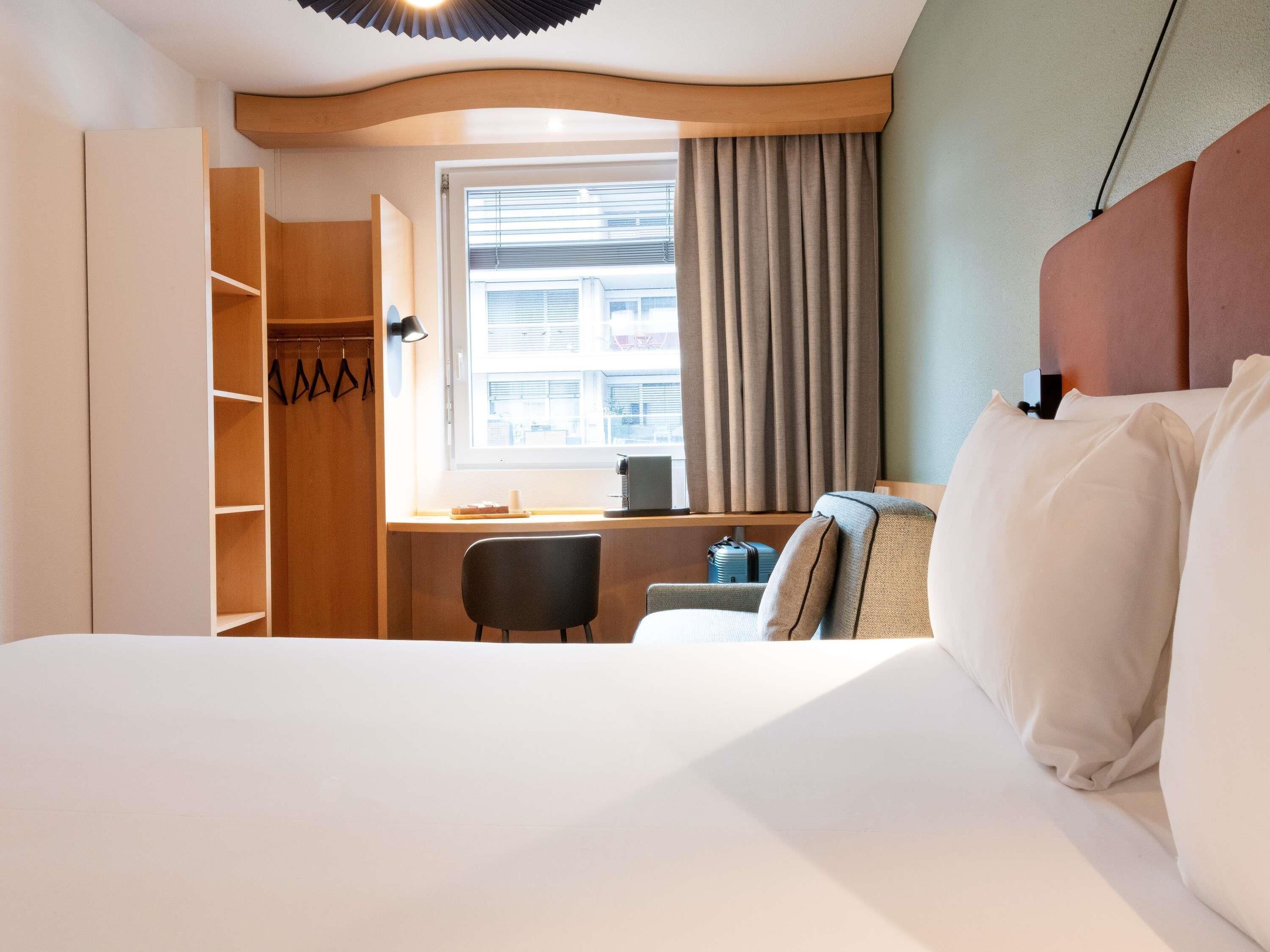 Habitació d'hotel amb un llit, una taula de treball, una cadira, un armari i una finestra amb cortines.