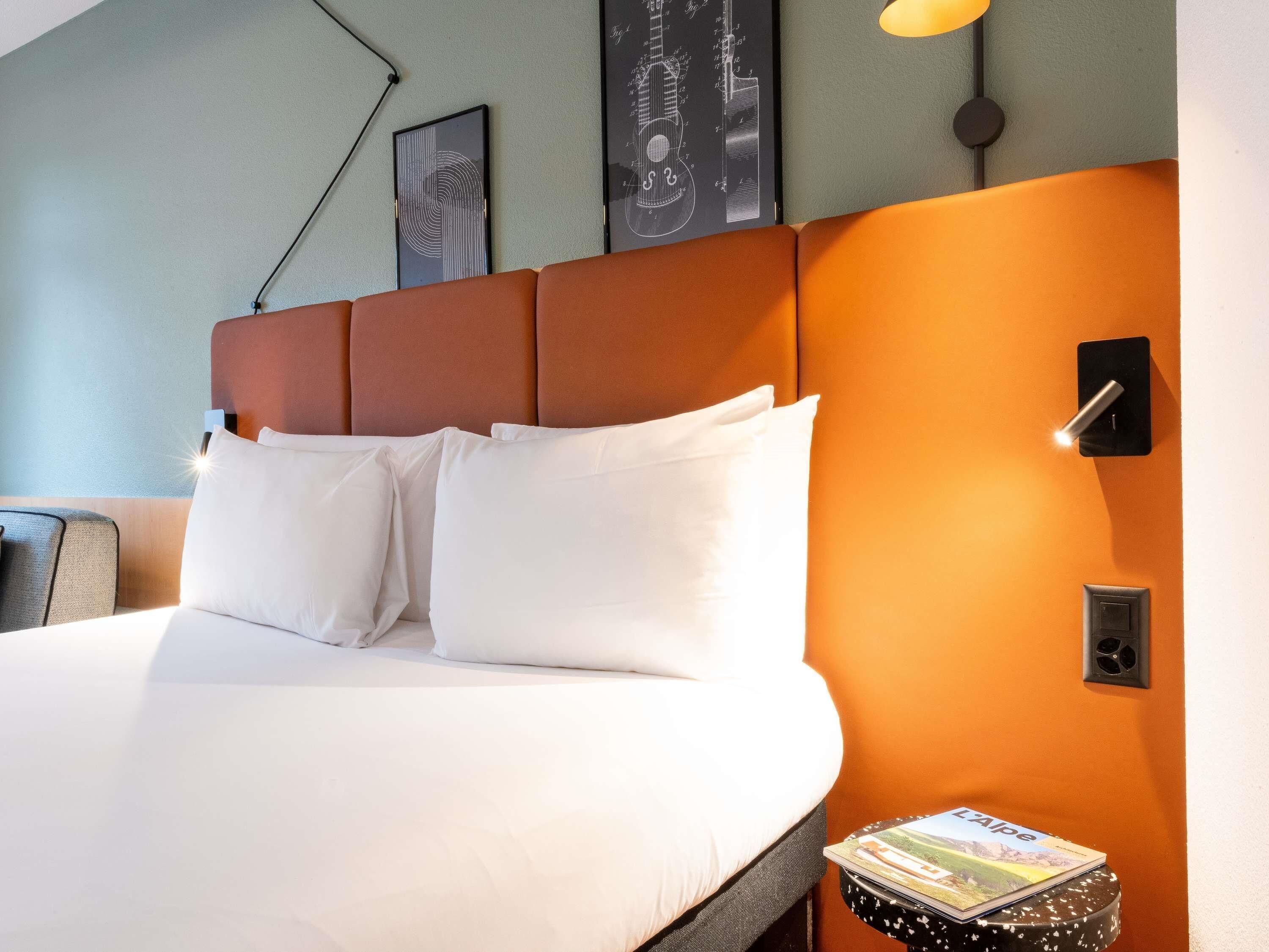 A hotel room with a large bed, white pillows, a headboard, a nightstand with a book, and wall art.