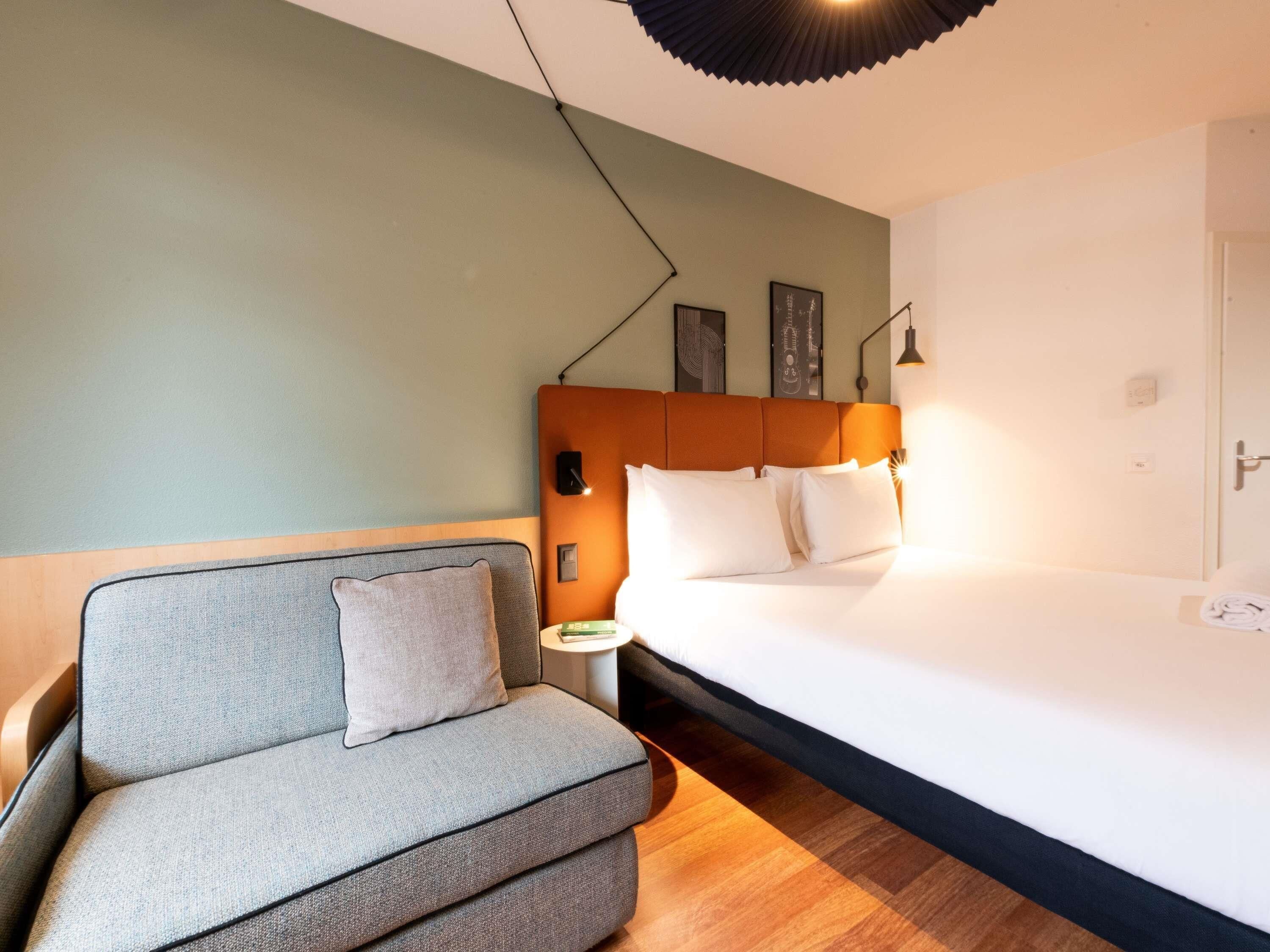 Una habitación de hotel moderna con un sofá gris, una cama grande con ropa blanca, piso de madera y una lámpara fijada a la pared.