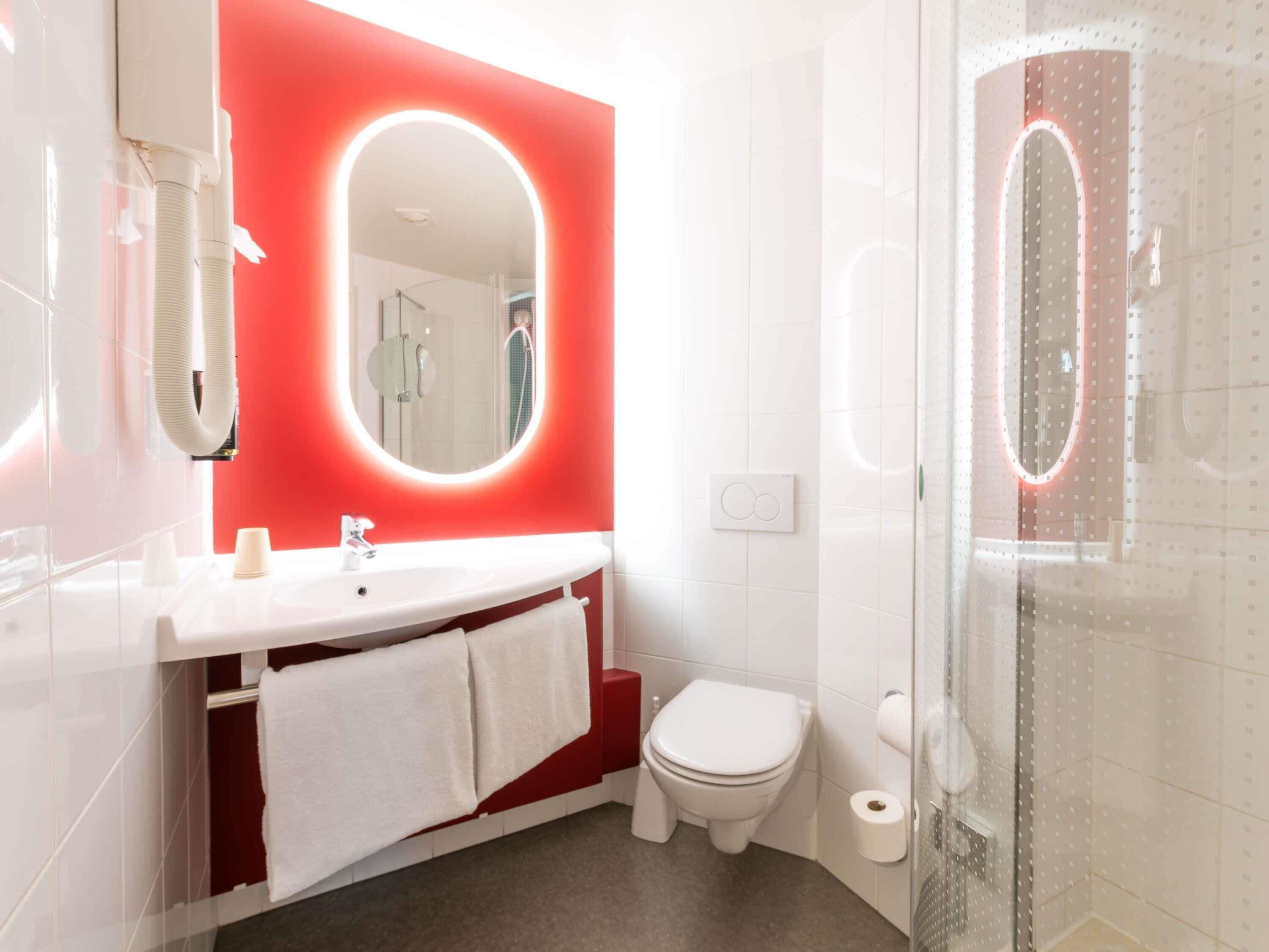 A modern bathroom with a white sink, a red accent wall, a mirror with a light, a toilet, and a shower area.