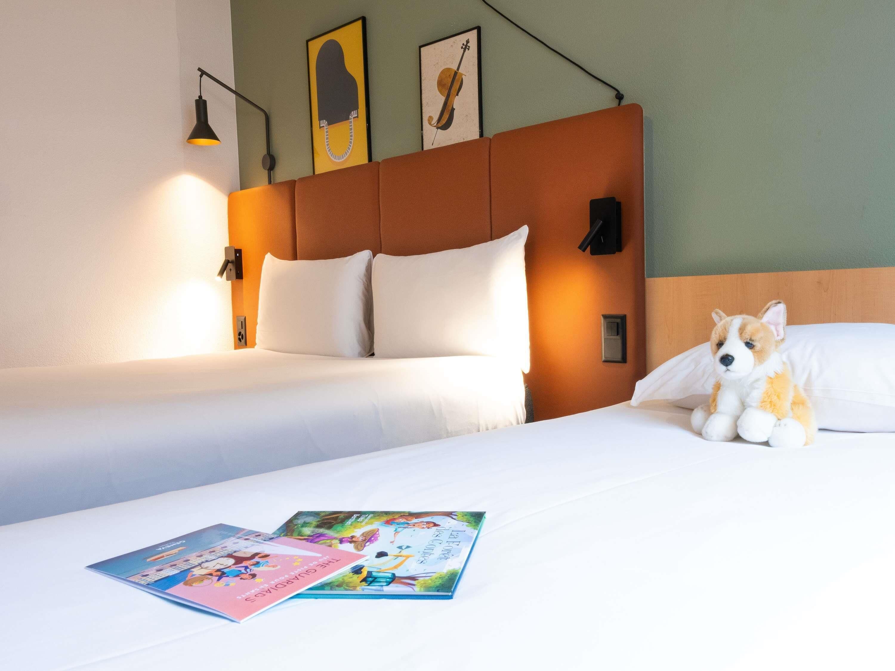 Habitación de hotel con dos camas, cabecero, lámparas fijadas a la pared y un peluche sobre la cama.