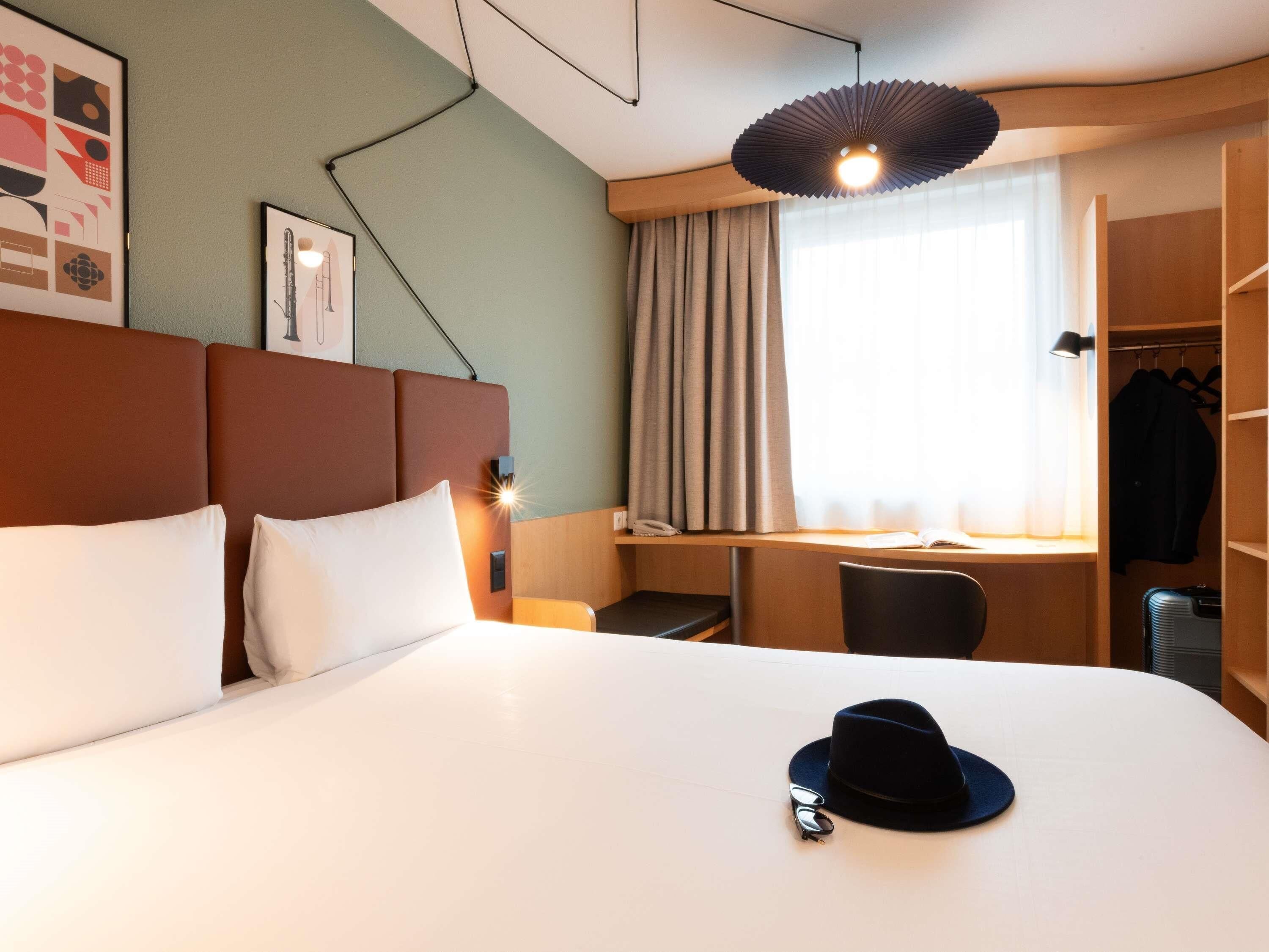 Habitación de hotel con cama, escritorio, silla, armario y lámpara colgante.
