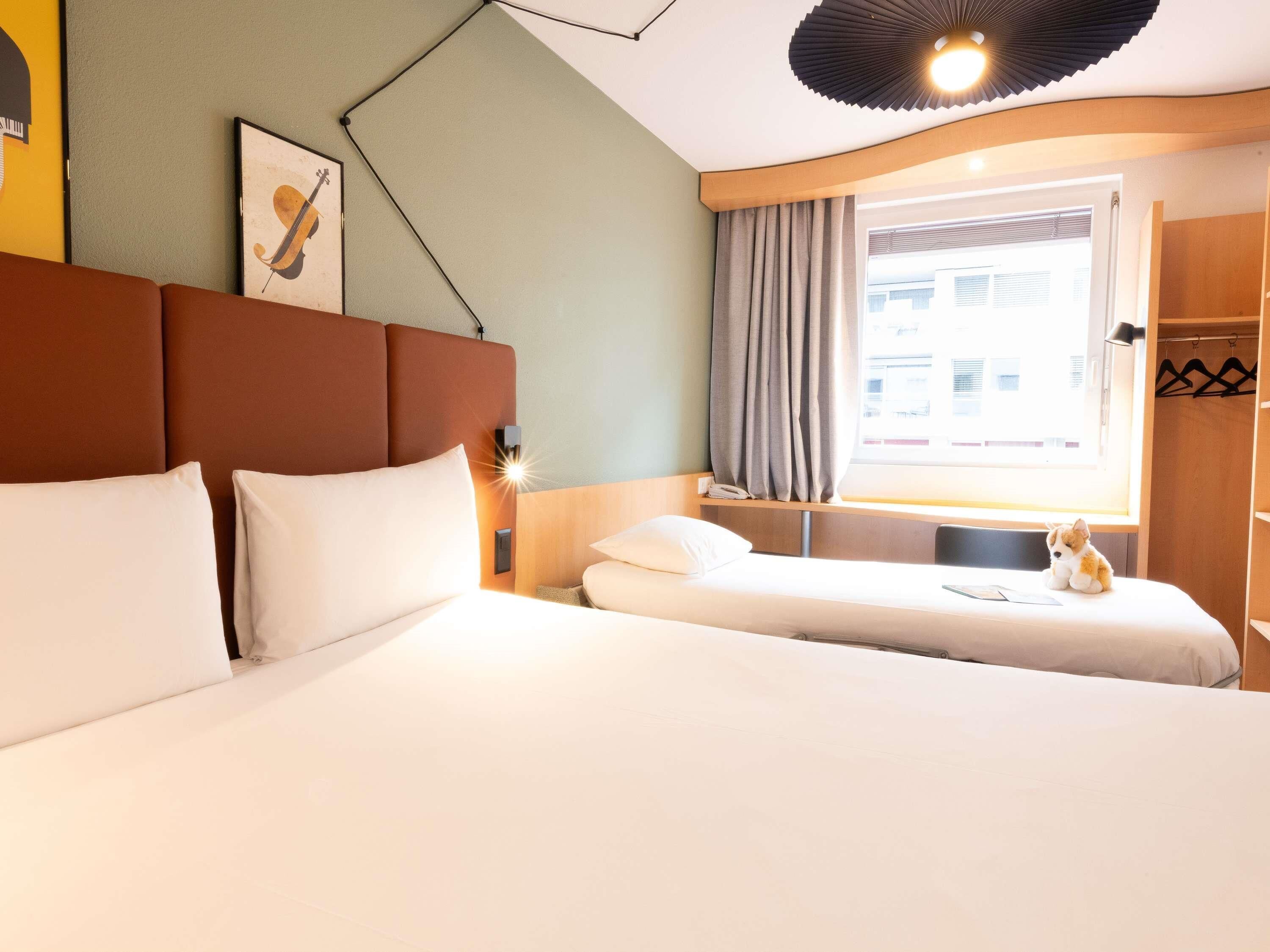 A hotel room with a large bed, a smaller bed, a desk with a lamp, a window with curtains, and a wall-mounted phone.