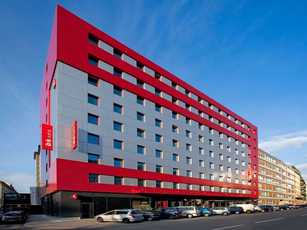 Un hotel moderno con fachada roja y gris, varios pisos y una fila de coches estacionados al frente.