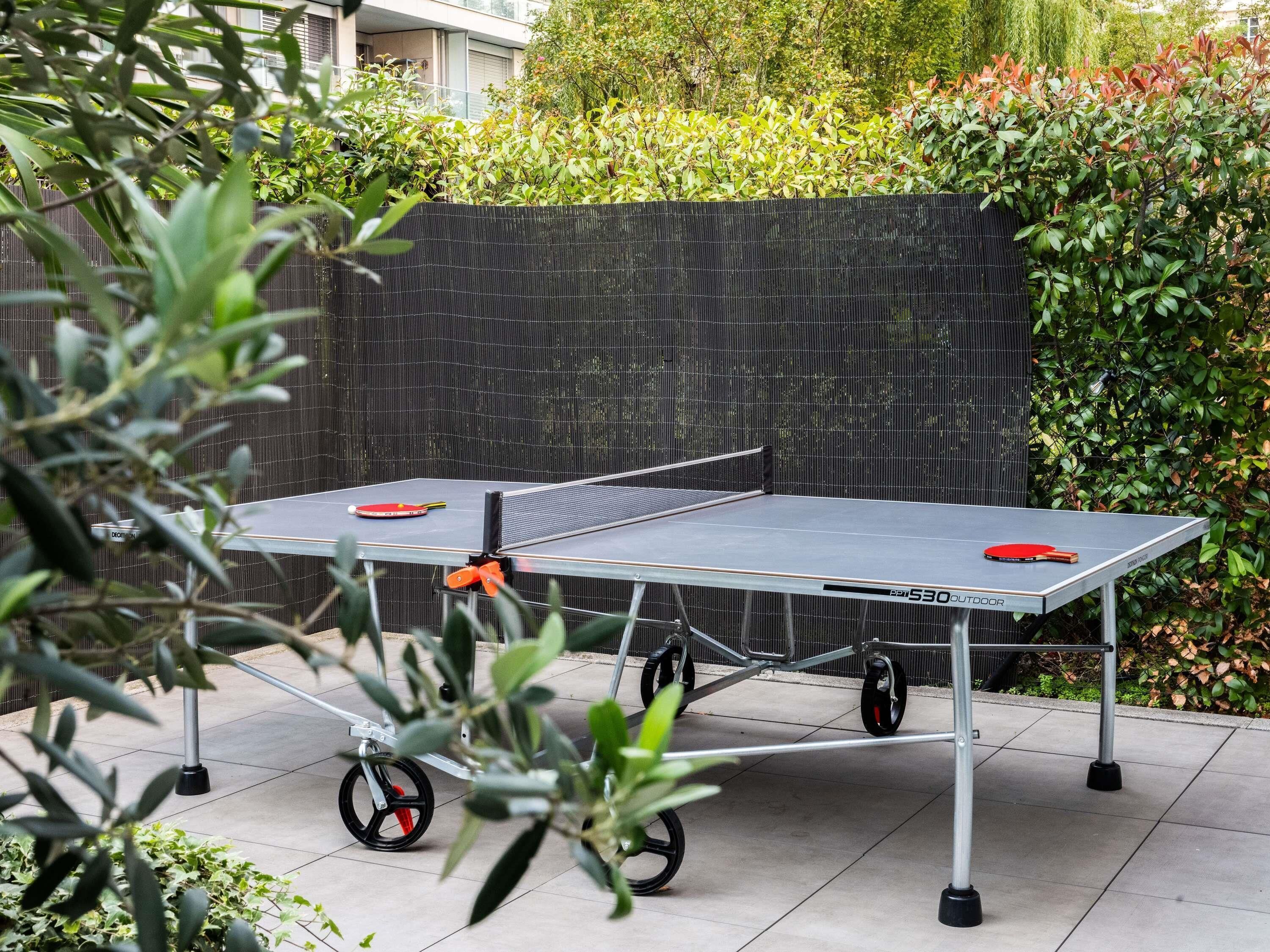 A portable ping pong table with wheels, set up outdoors with a black mesh backdrop and greenery in the background.