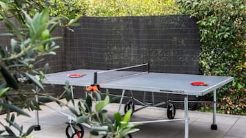 Una mesa de ping pong portátil con ruedas, montada al aire libre con un fondo de malla negra y follaje en el fondo.