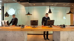 A reception desk with two employees, a modern interior design, and a projection screen displaying a menu. - ibis Geneve Centre Nations (Geneva)