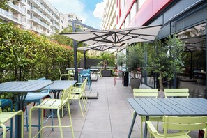 Um pátio externo com mesas e cadeiras, cercado por vegetação e edifícios.