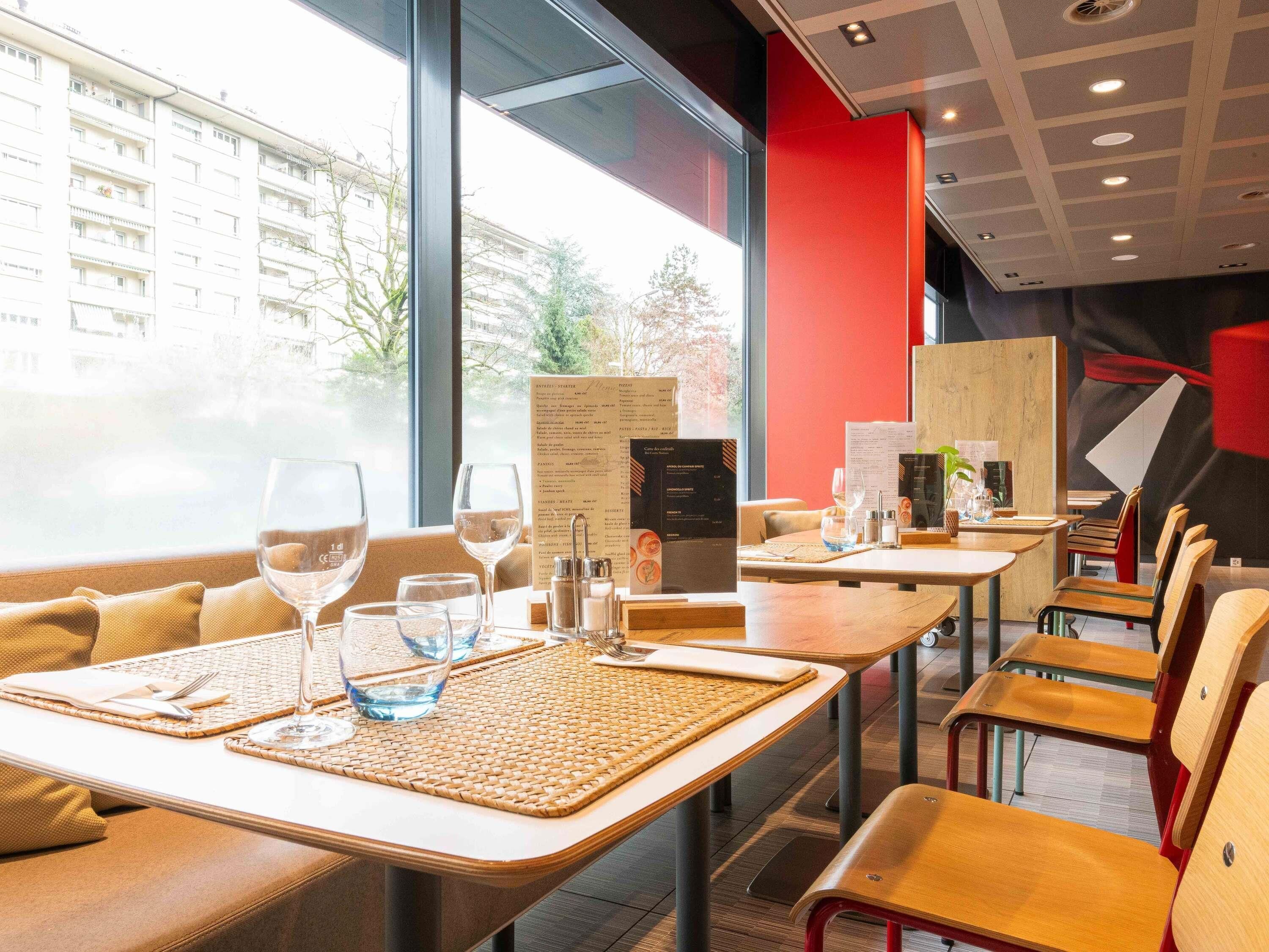 Un restaurant où les tables sont dressées pour le service, où les menus sont posés sur les tables et d’où l’on peut admirer les bâtiments environnants.