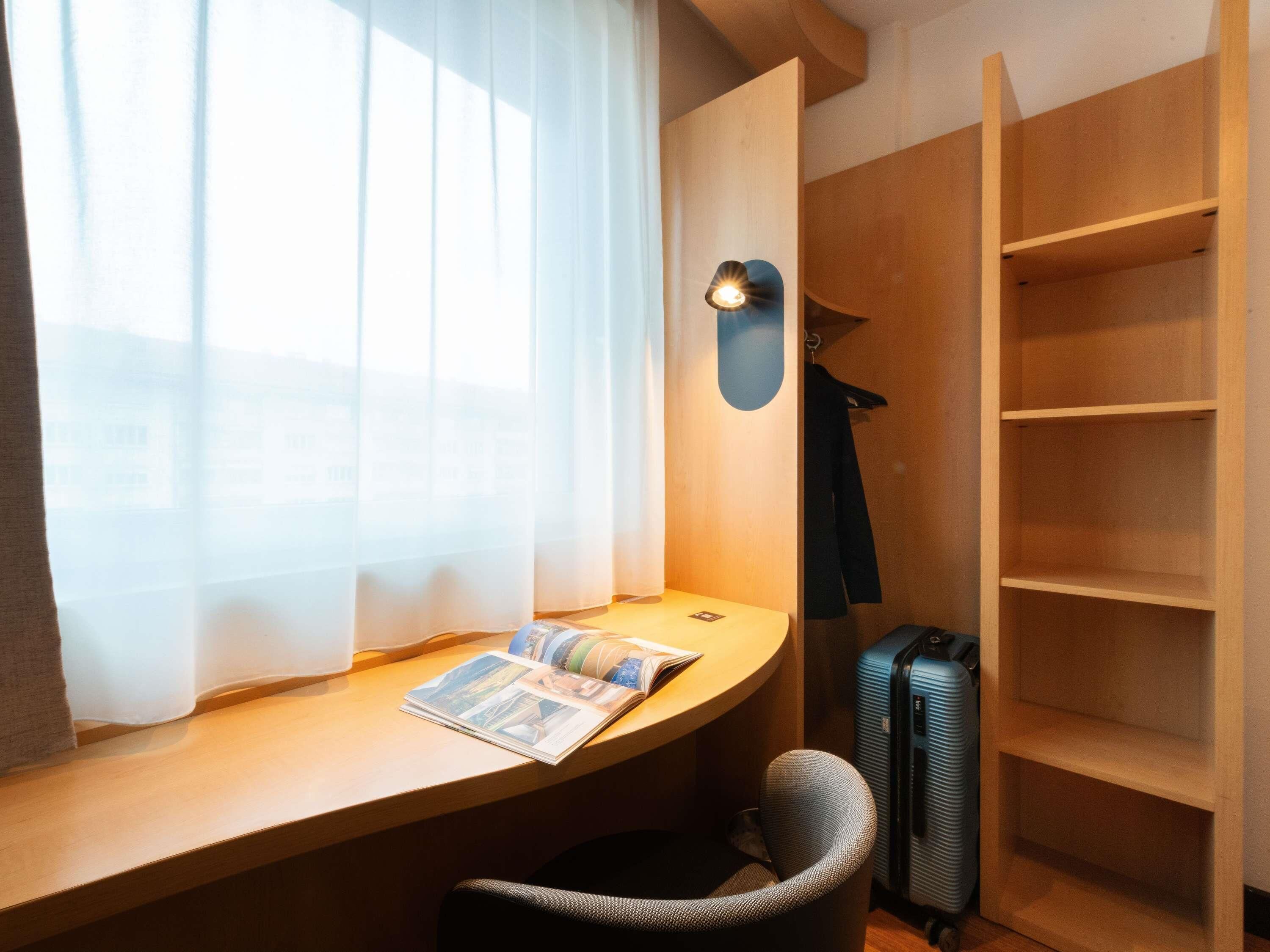 Quarto de hotel com escrivaninha, cadeira, luminária, mala e guarda-roupa.